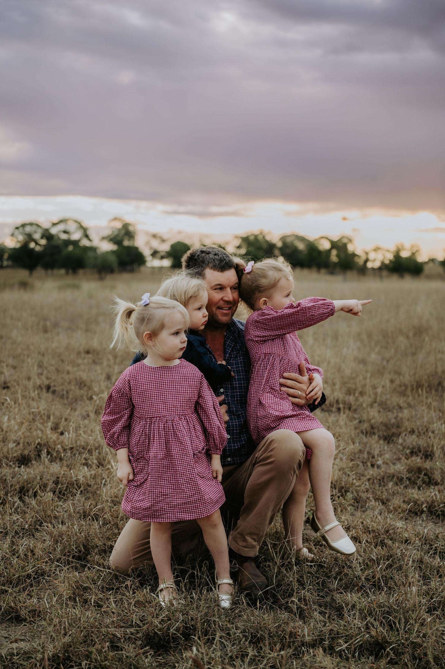 A father crouches in a field at sunset with two young girls in matching pink dresses, one pointing into the distance.
