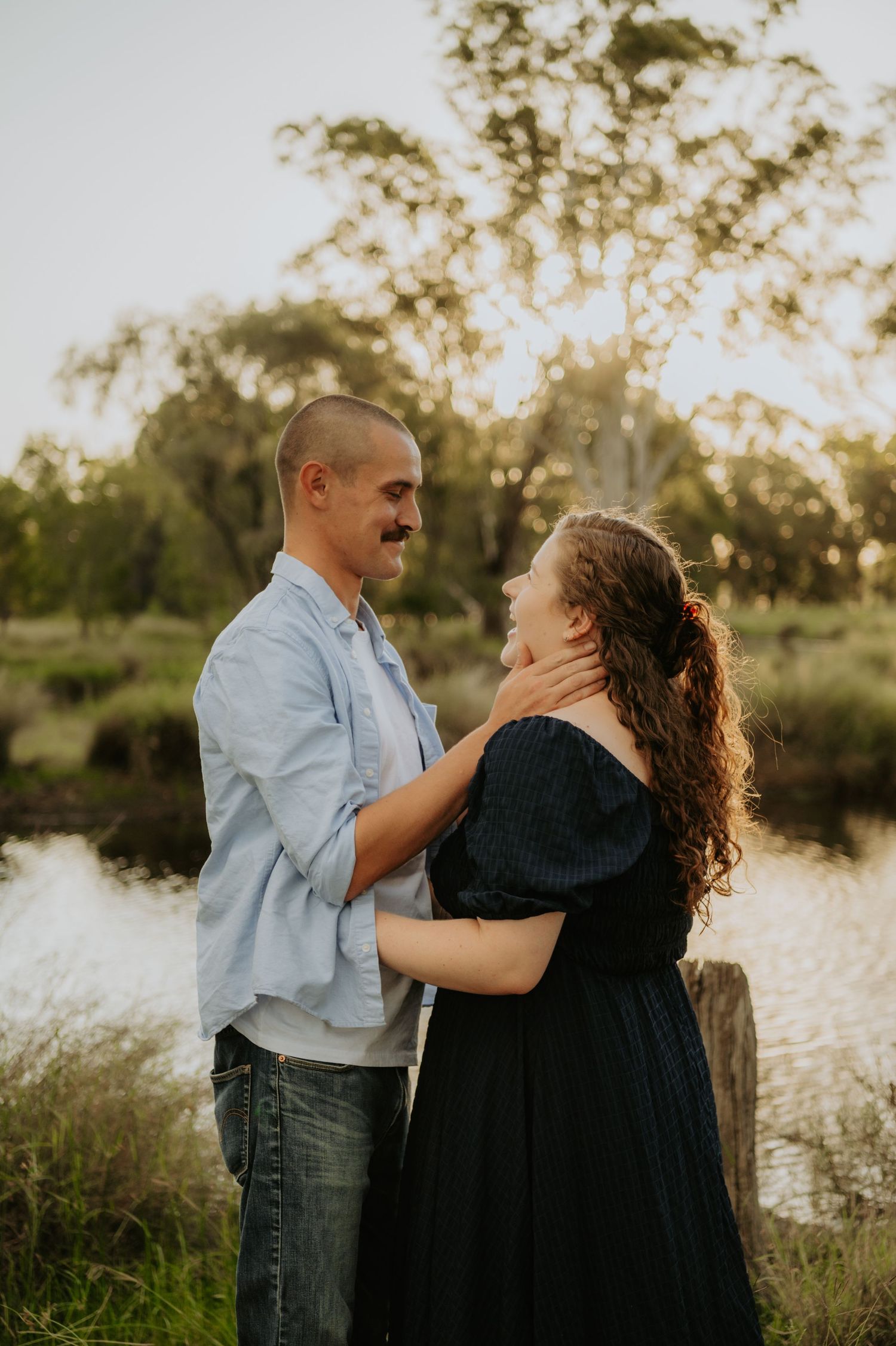 Couple sharing a romantic moment outdoors near a pond, man in blue shirt gently holding woman's face in golden hour light.