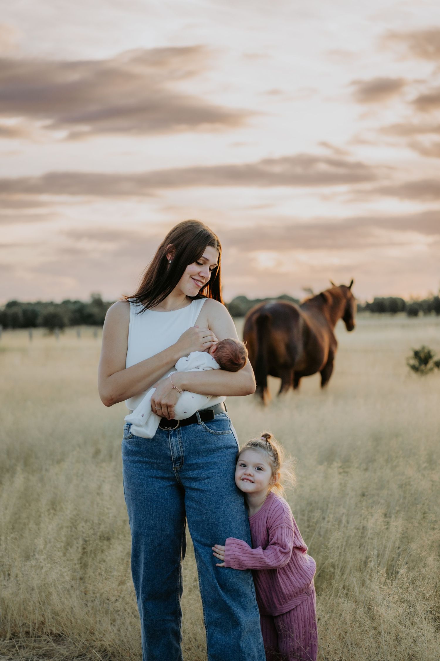 A mother holds a newborn while her young daughter hugs her leg in a golden field at sunset with a horse nearby.