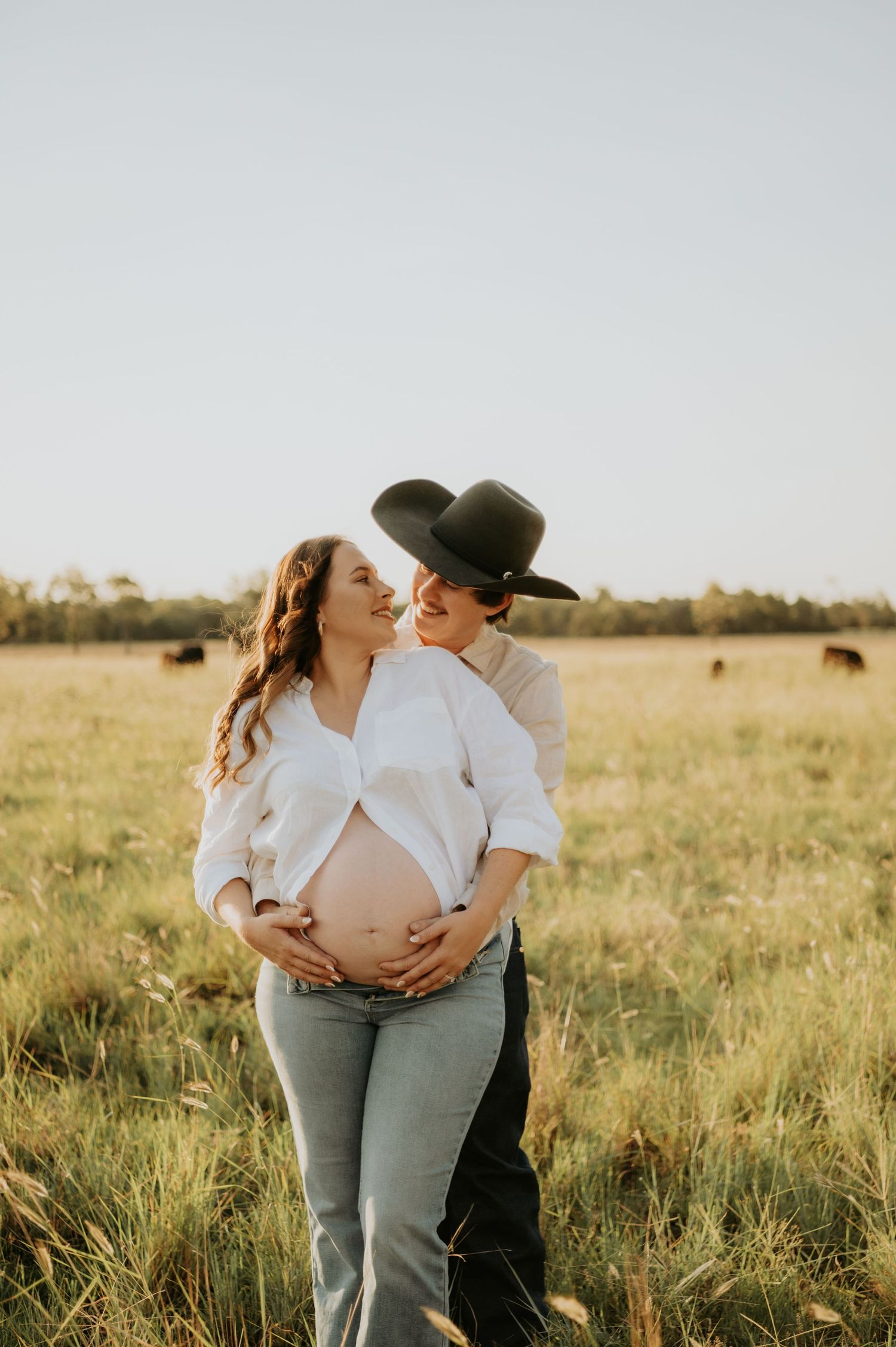 A couple shares an intimate moment in a sunlit field during a maternity photoshoot at golden hour.