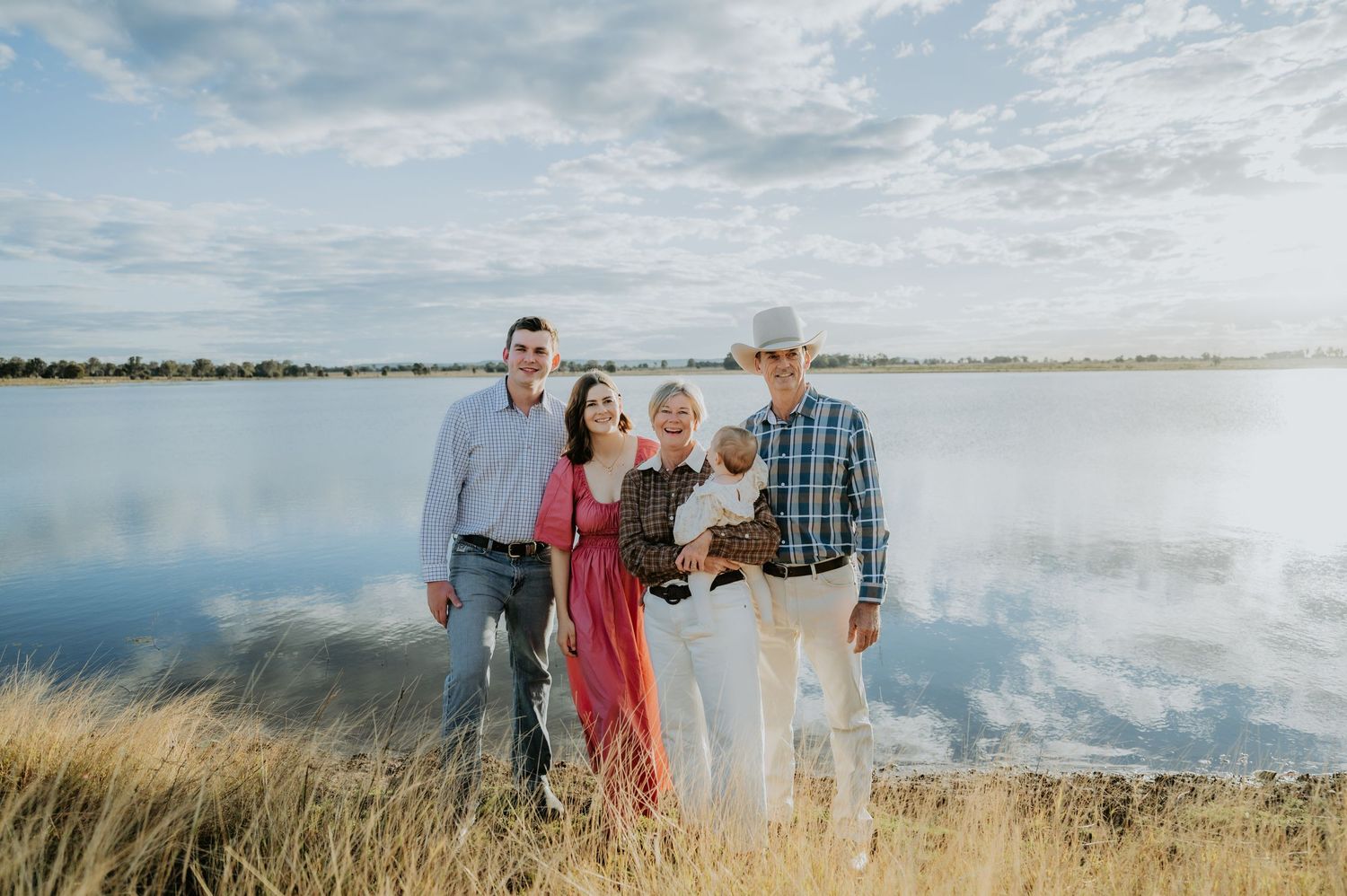 Family of five posing together near a reflective lake with golden grass and cloudy sky backdrop.