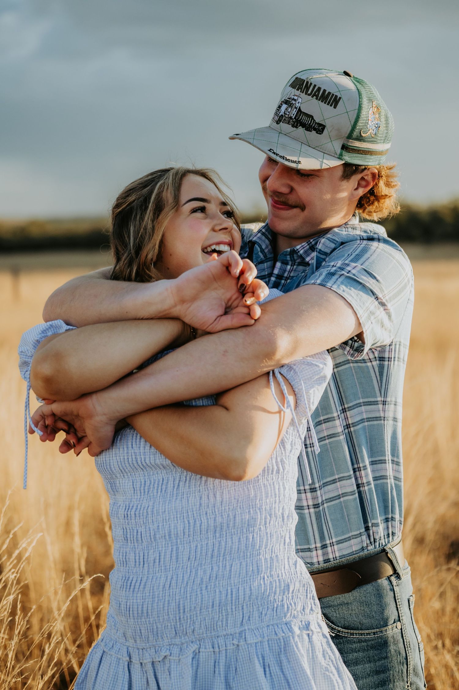 A couple embraces in a golden wheat field, the man in a plaid shirt and cap hugging a smiling woman in a white dress.