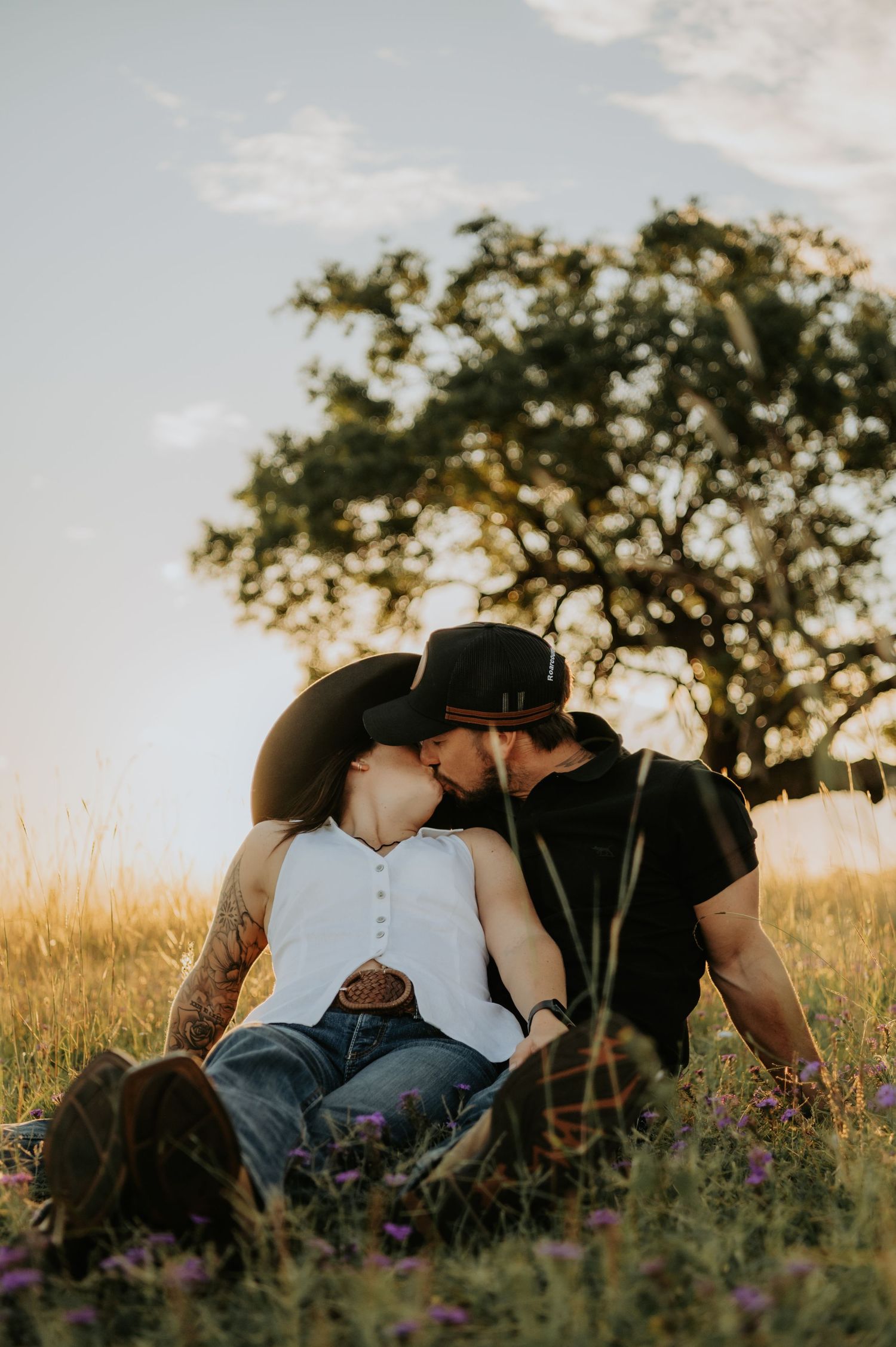 Couple kissing in a golden field at sunset, wearing cowboy hats, sitting among wildflowers with a tree in the background.