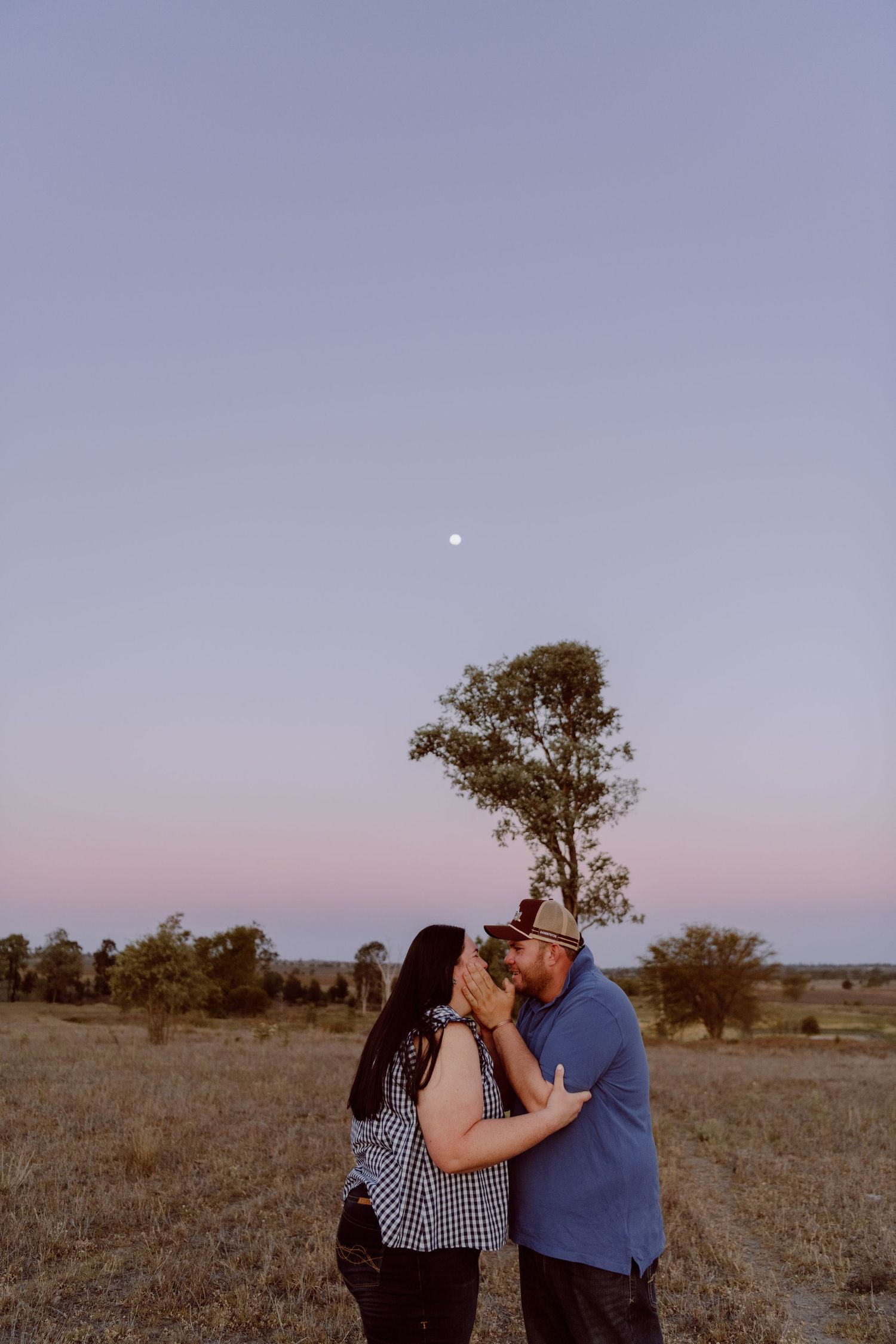 Couple sharing a romantic kiss in an open field at dusk with a lone tree and moon visible in the pastel sky.