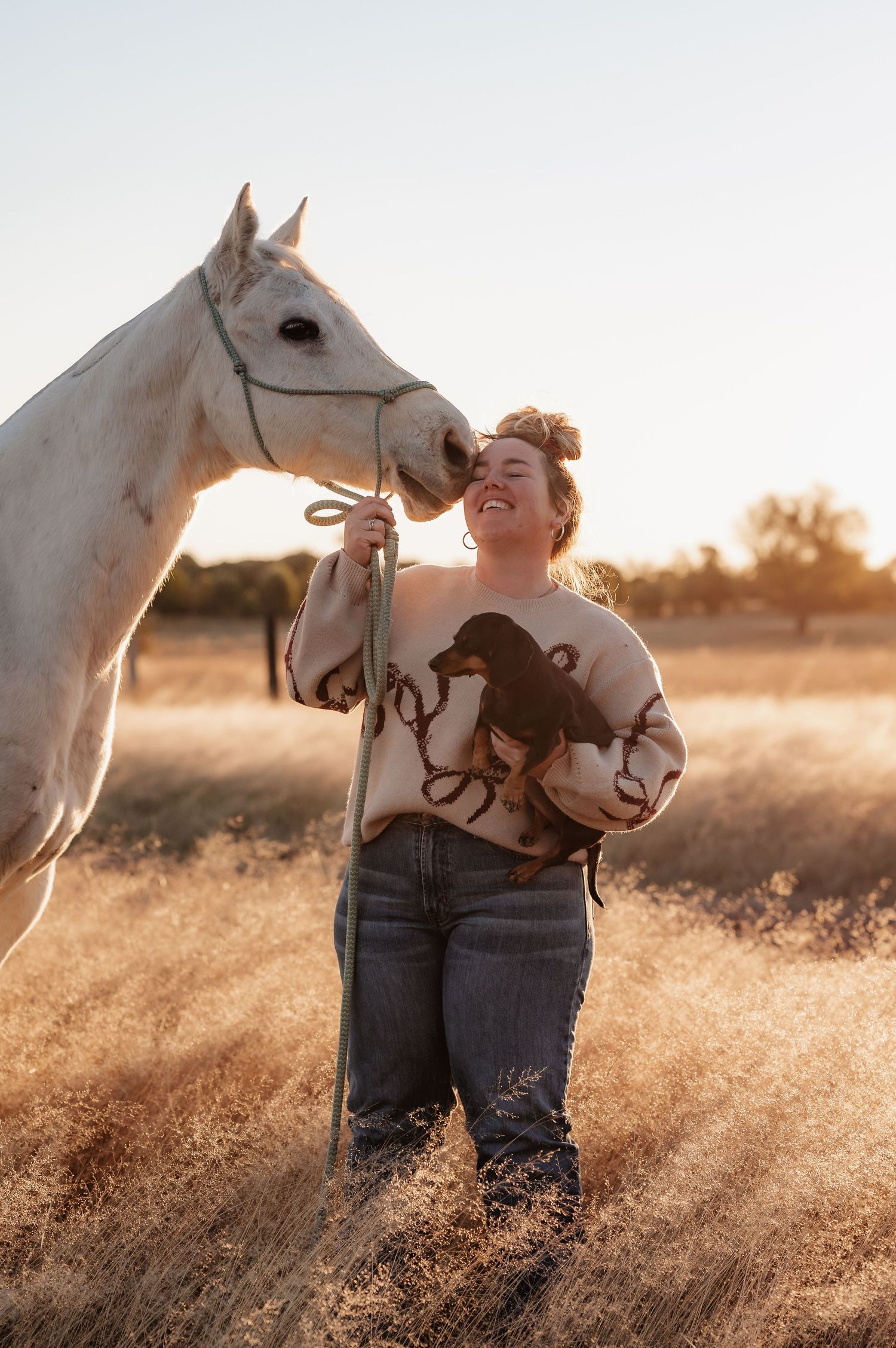 Woman laughing with white horse in golden field at sunset, wearing dachshund sweater and jeans.