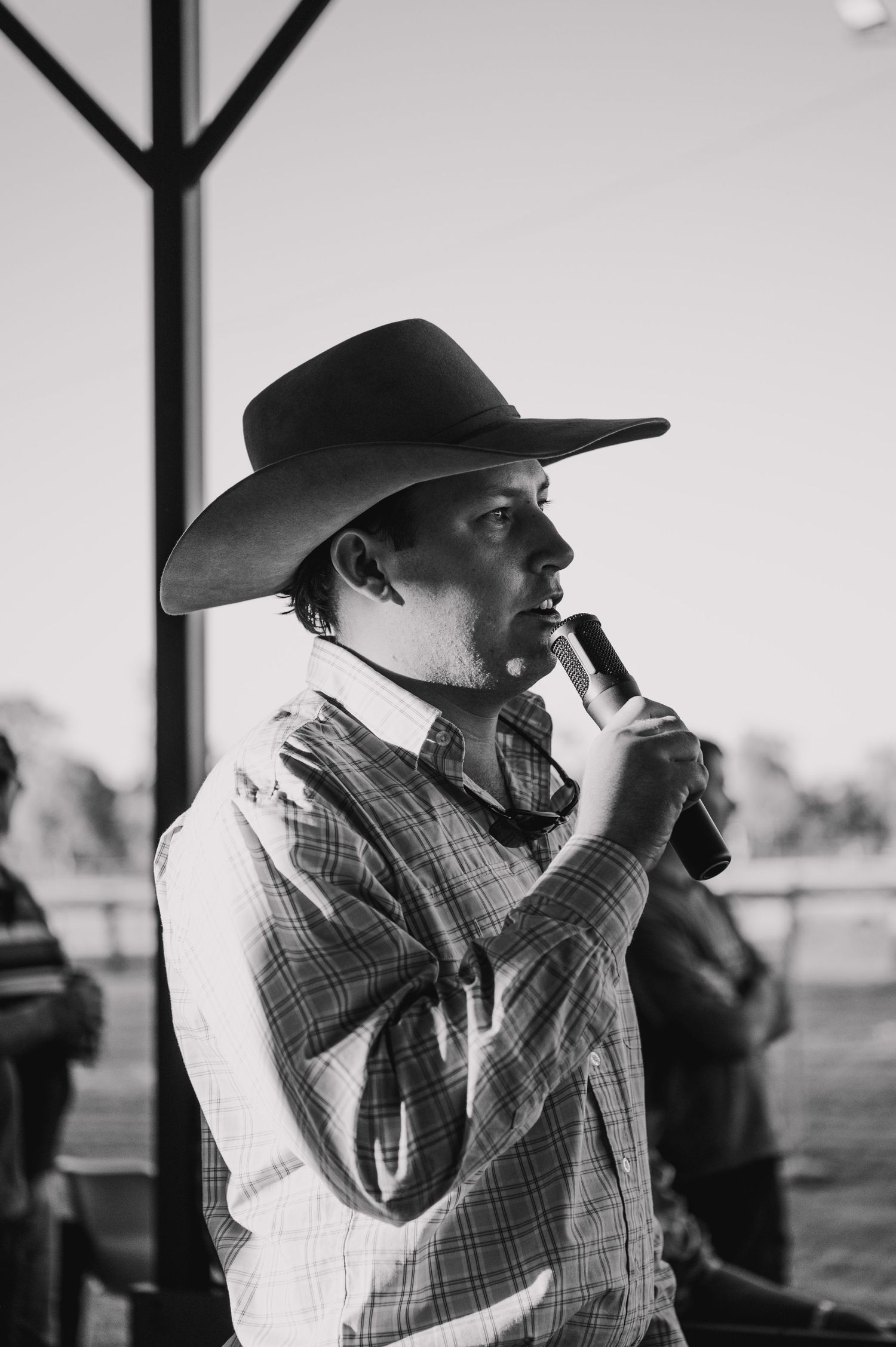 Black and white photo of a man in a cowboy hat holding a microphone outdoors at a rodeo event.