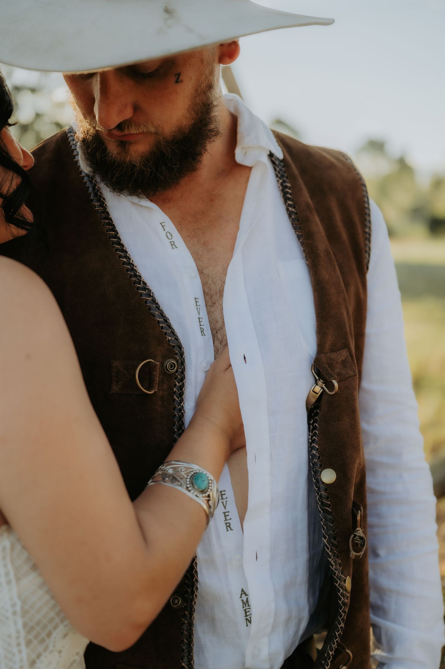 Bearded man in white shirt, brown leather vest, and cowboy hat embraces a woman outdoors.