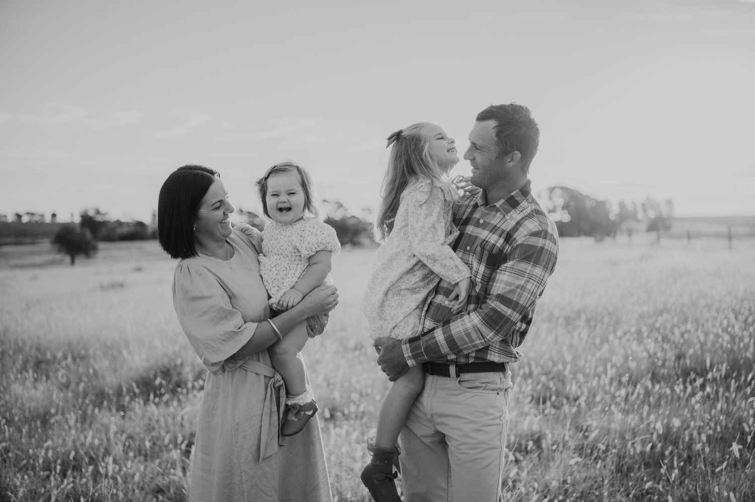 Black and white photo of a happy family of four laughing together in a sunlit field.