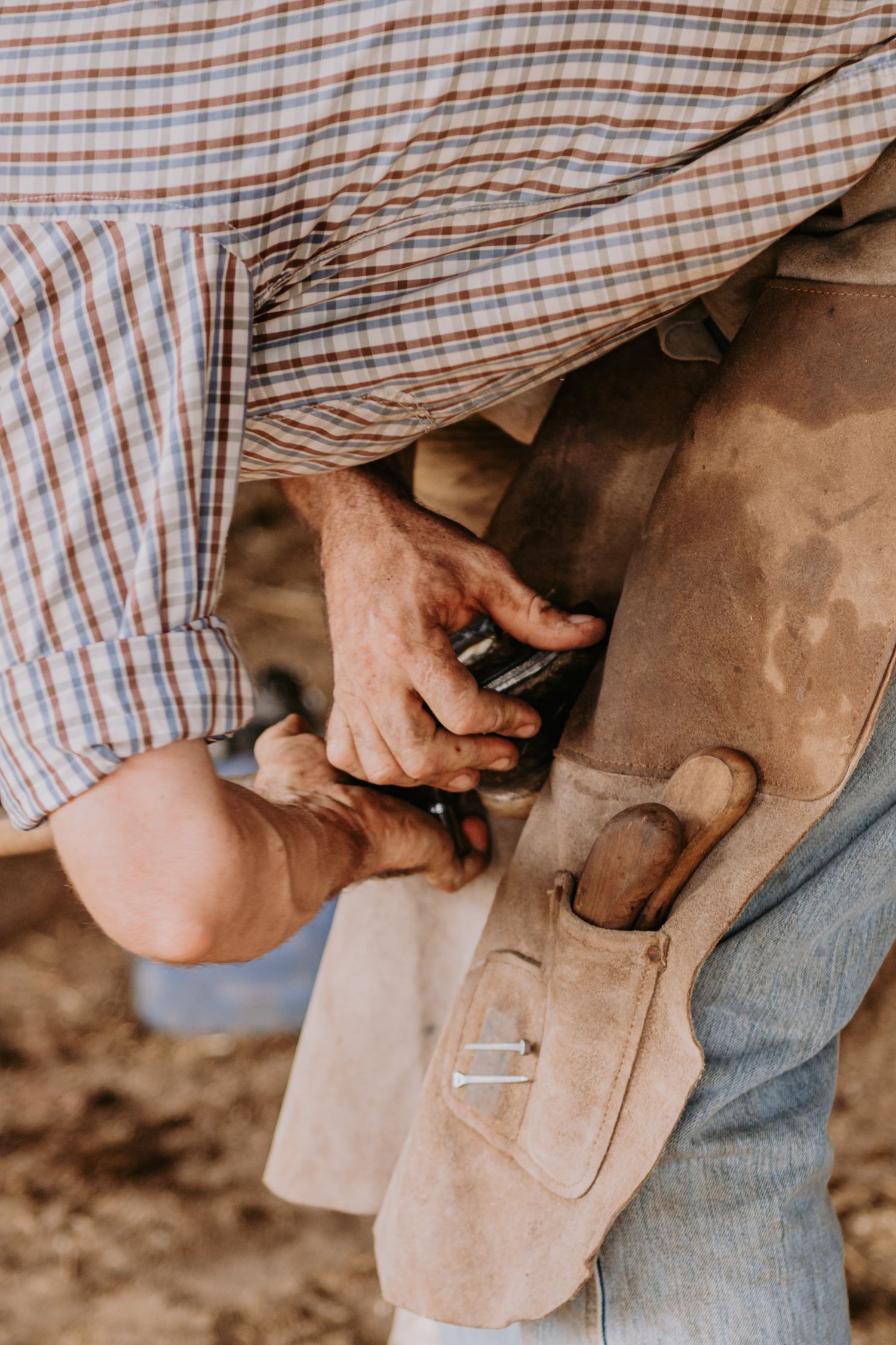 A farrier in a plaid shirt uses tools to shoe a horse hoof, wearing a leather apron outdoors.