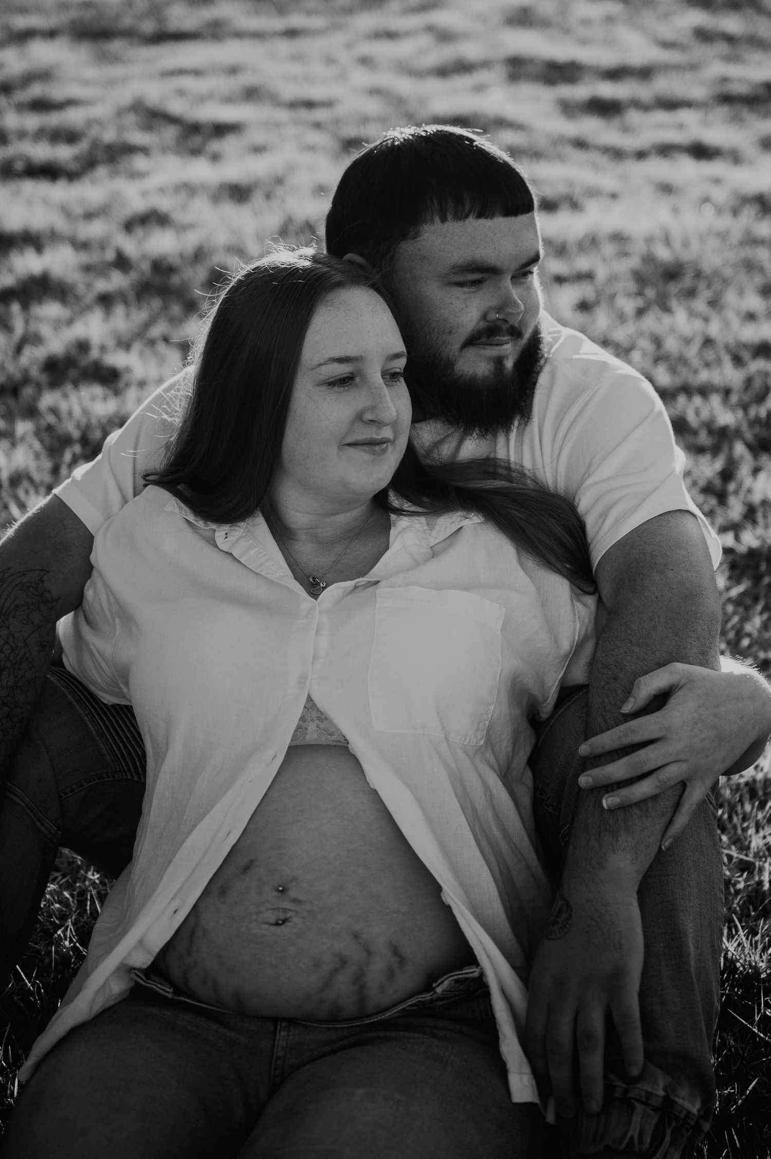 Black and white maternity photo of a pregnant woman and her partner sitting together in a grassy field.