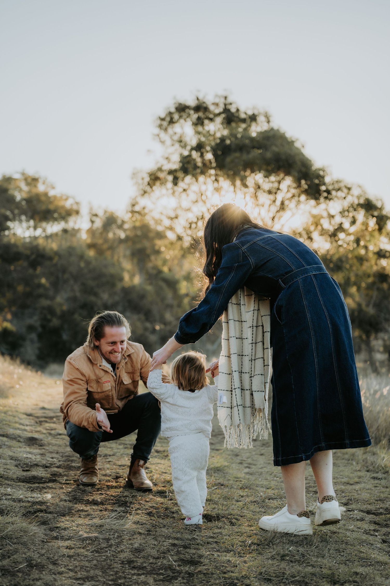 A young family helps their toddler take first steps outdoors in golden hour light on a grassy path.