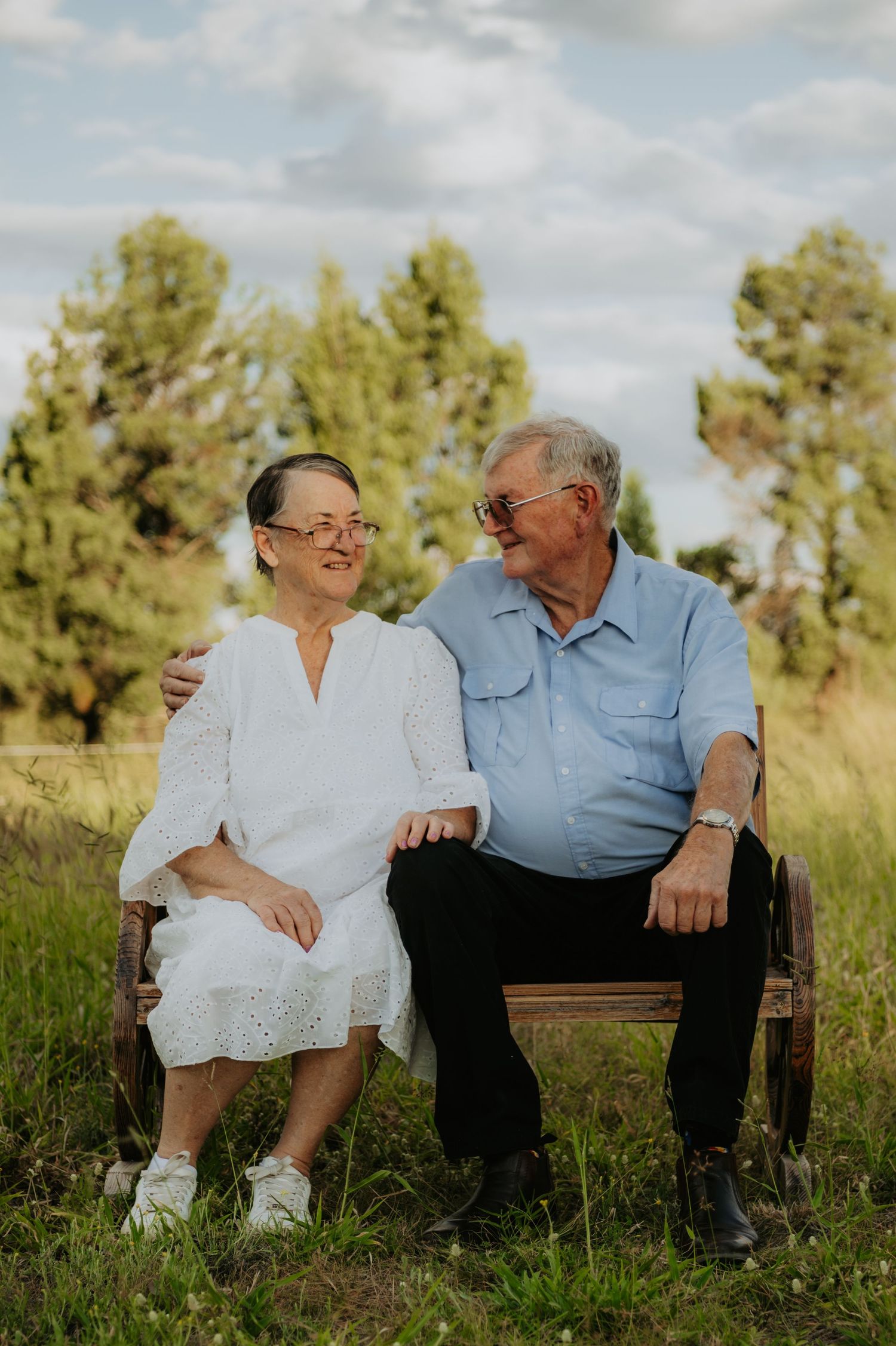 Elderly couple sitting on a wooden bench outdoors, smiling at each other, surrounded by green trees and grass.