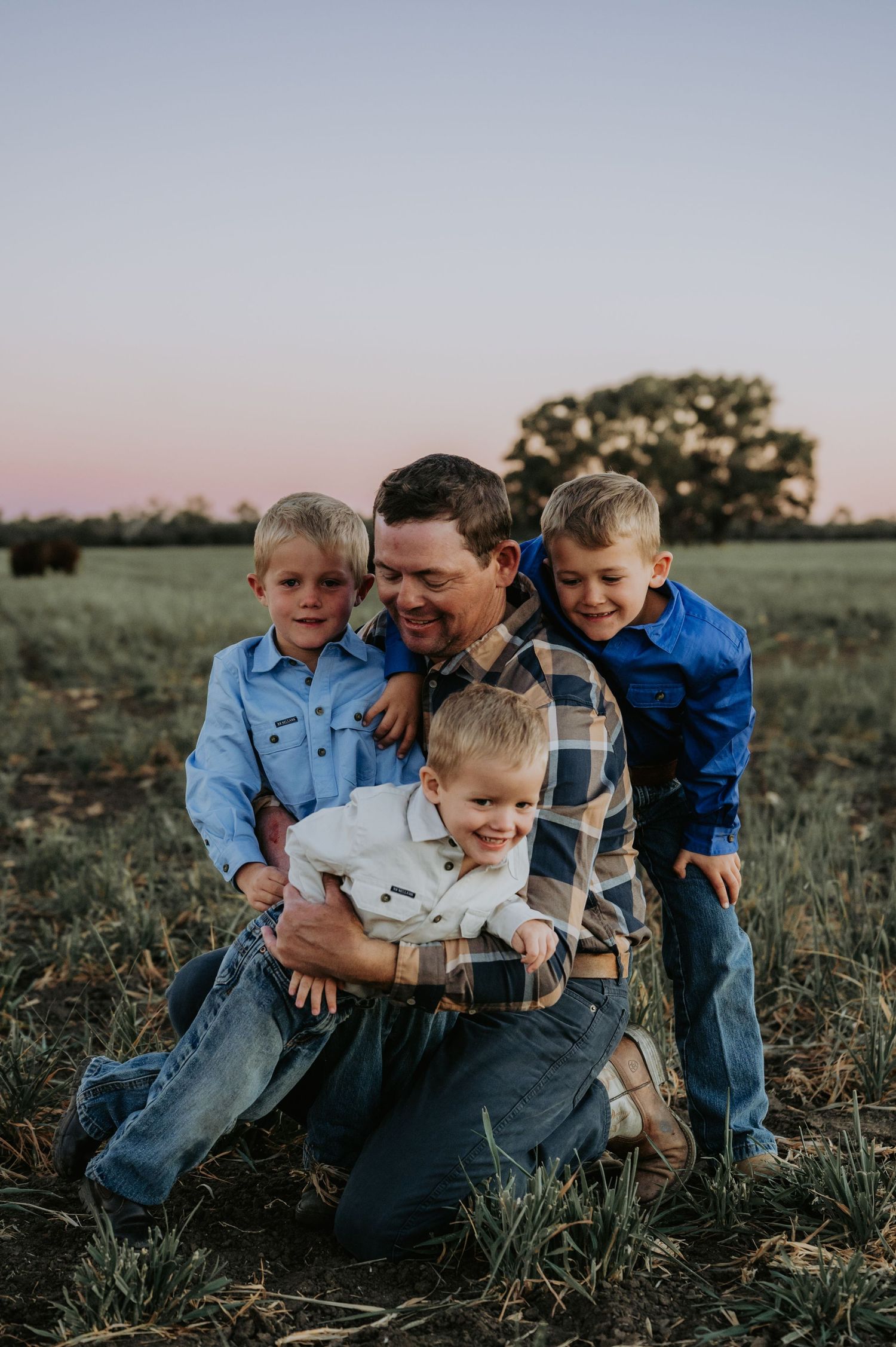 A father kneels in a field at sunset, joyfully embracing his three young sons dressed in blue shirts and jeans.
