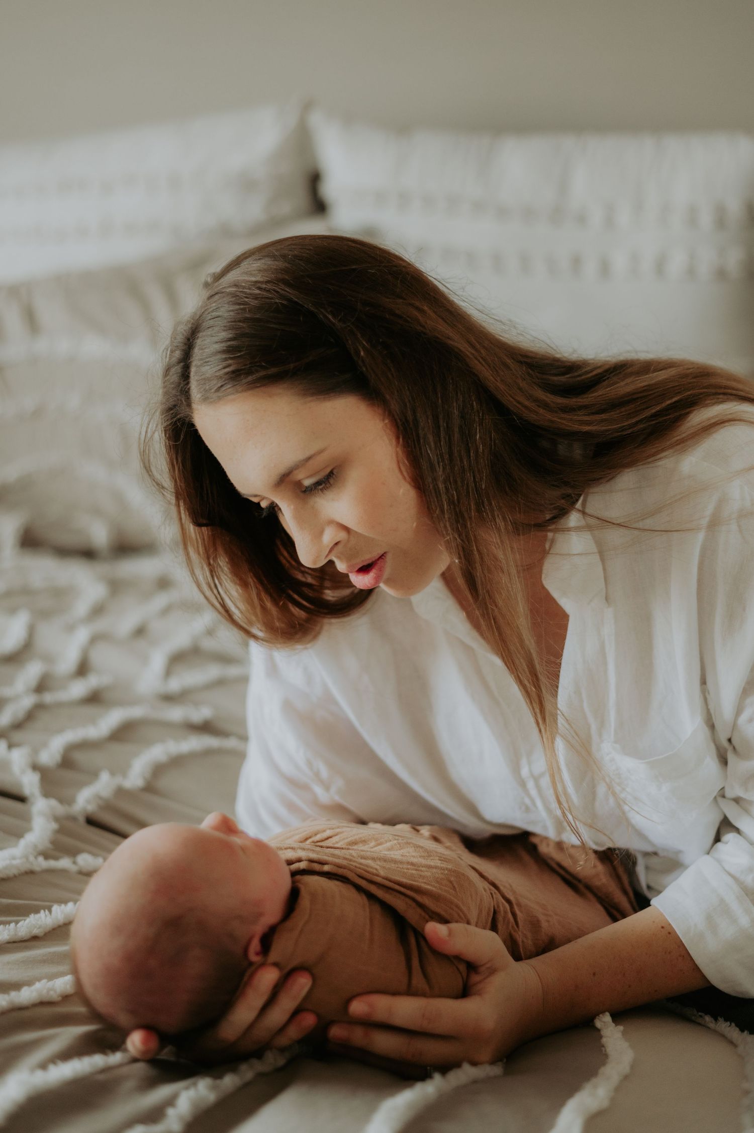 Mother in white dress lying on bed tenderly nursing and bonding with baby in soft natural lighting.