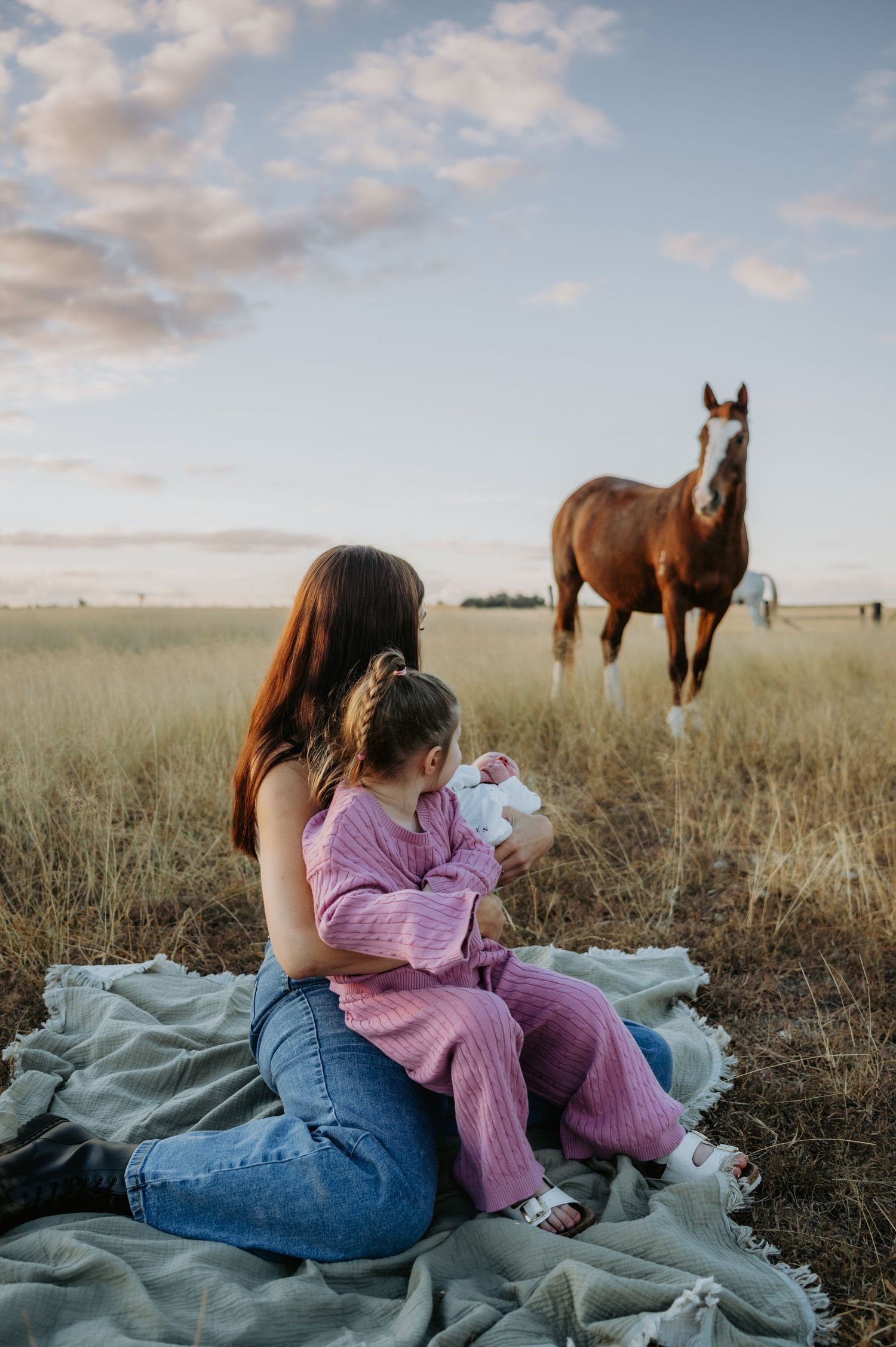 Mother and daughter sitting on blanket in field holding newborn baby with brown horse grazing in background.