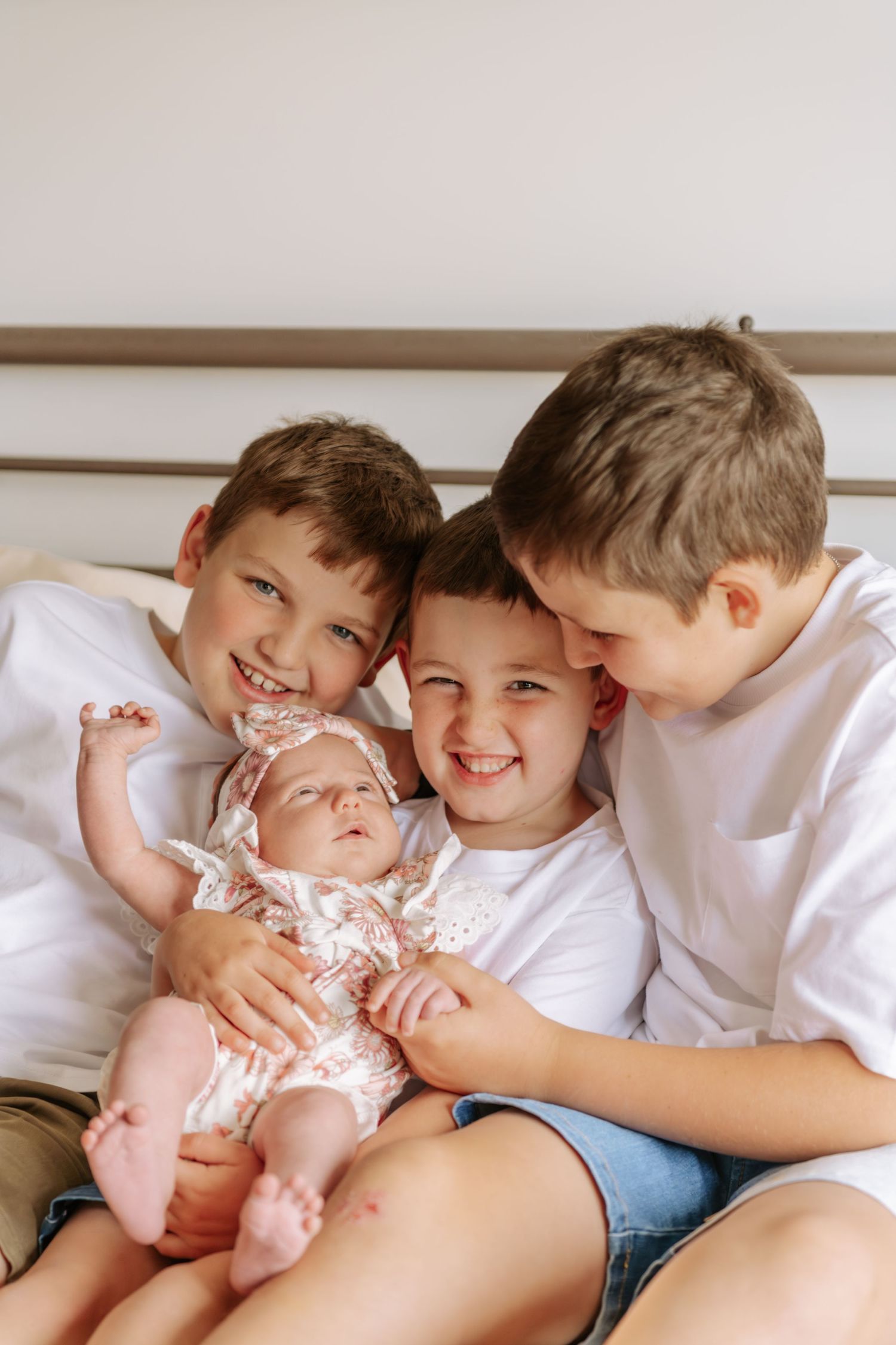 Three smiling boys in white shirts cuddle together on a bed while holding their newborn baby sister.