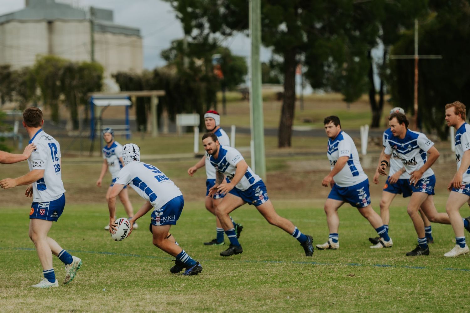Rugby league players in white and blue uniforms compete during an outdoor match on a green grass field.