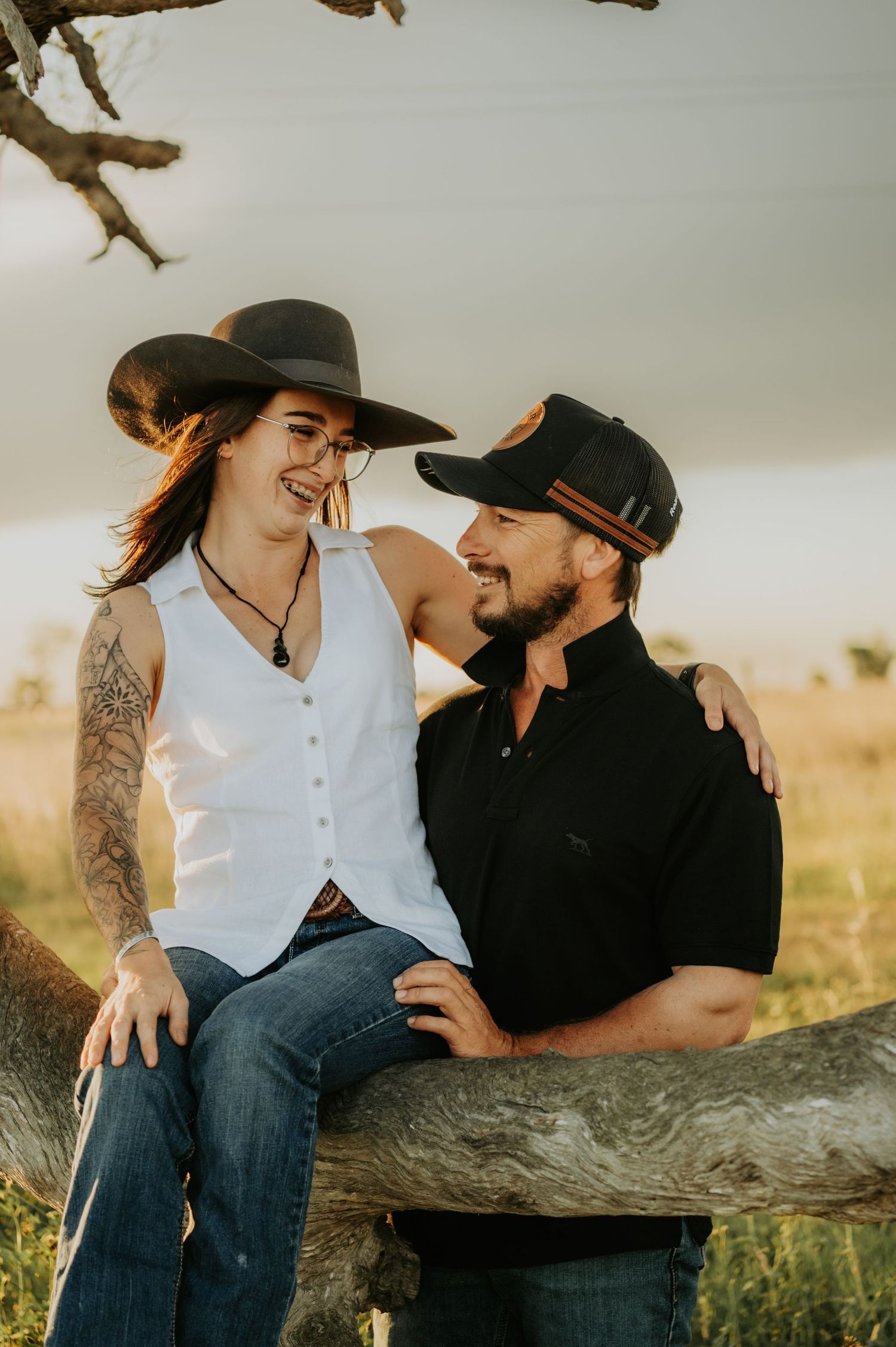 A romantic couple in western attire share intimate moments while sitting on a fallen tree trunk at sunset.
