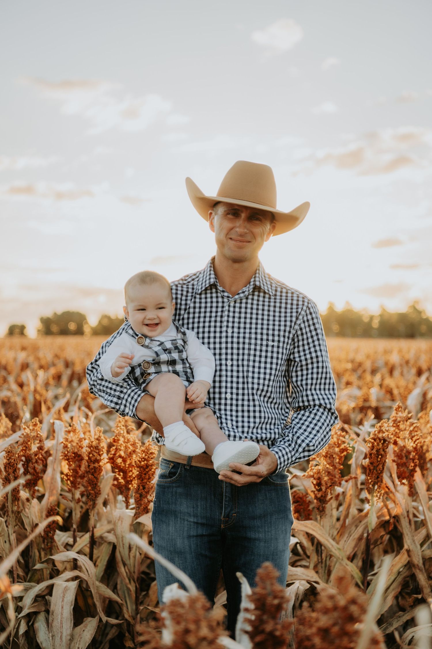 Man in cowboy hat holding smiling baby in a golden sorghum field at sunset.