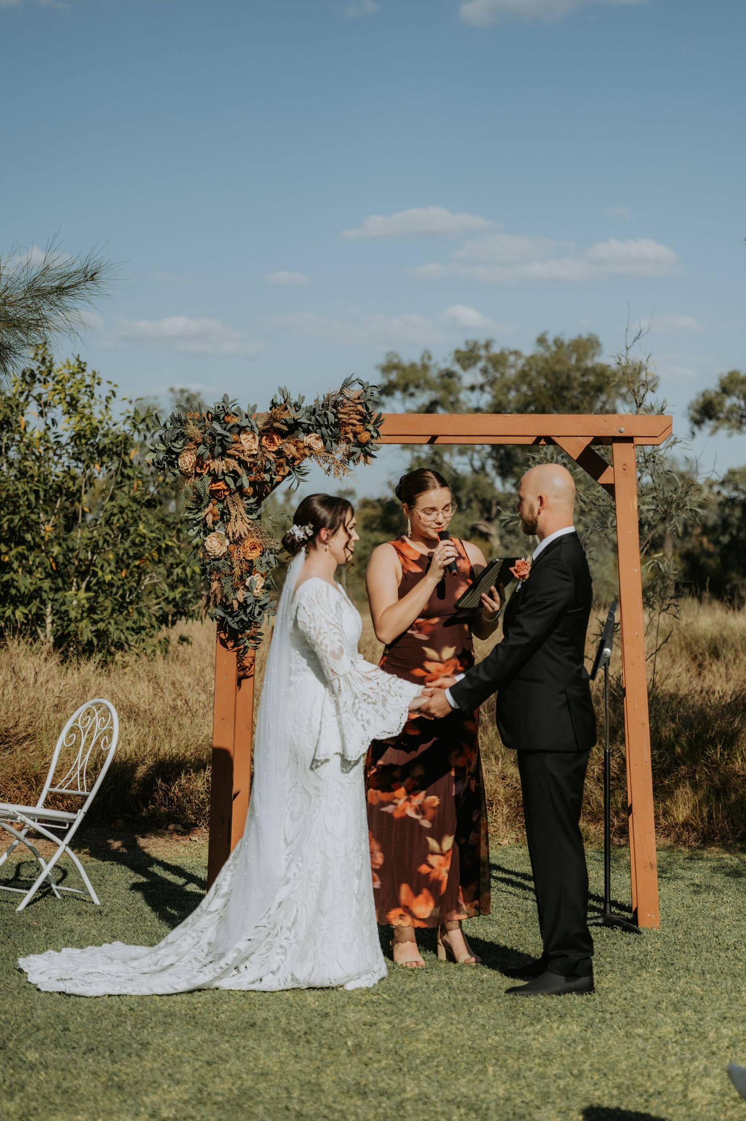 Outdoor wedding ceremony under wooden arch with bride in lace gown, groom in black suit, and officiant in floral dress.