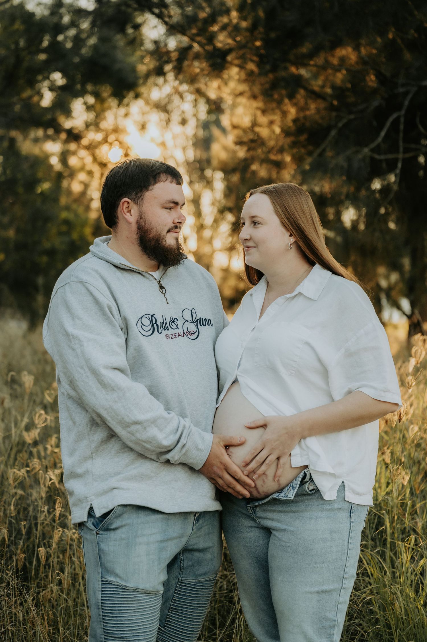A couple tenderly cradles a baby bump in a golden sunlit field, wearing casual jeans and sweatshirts for a maternity photo.