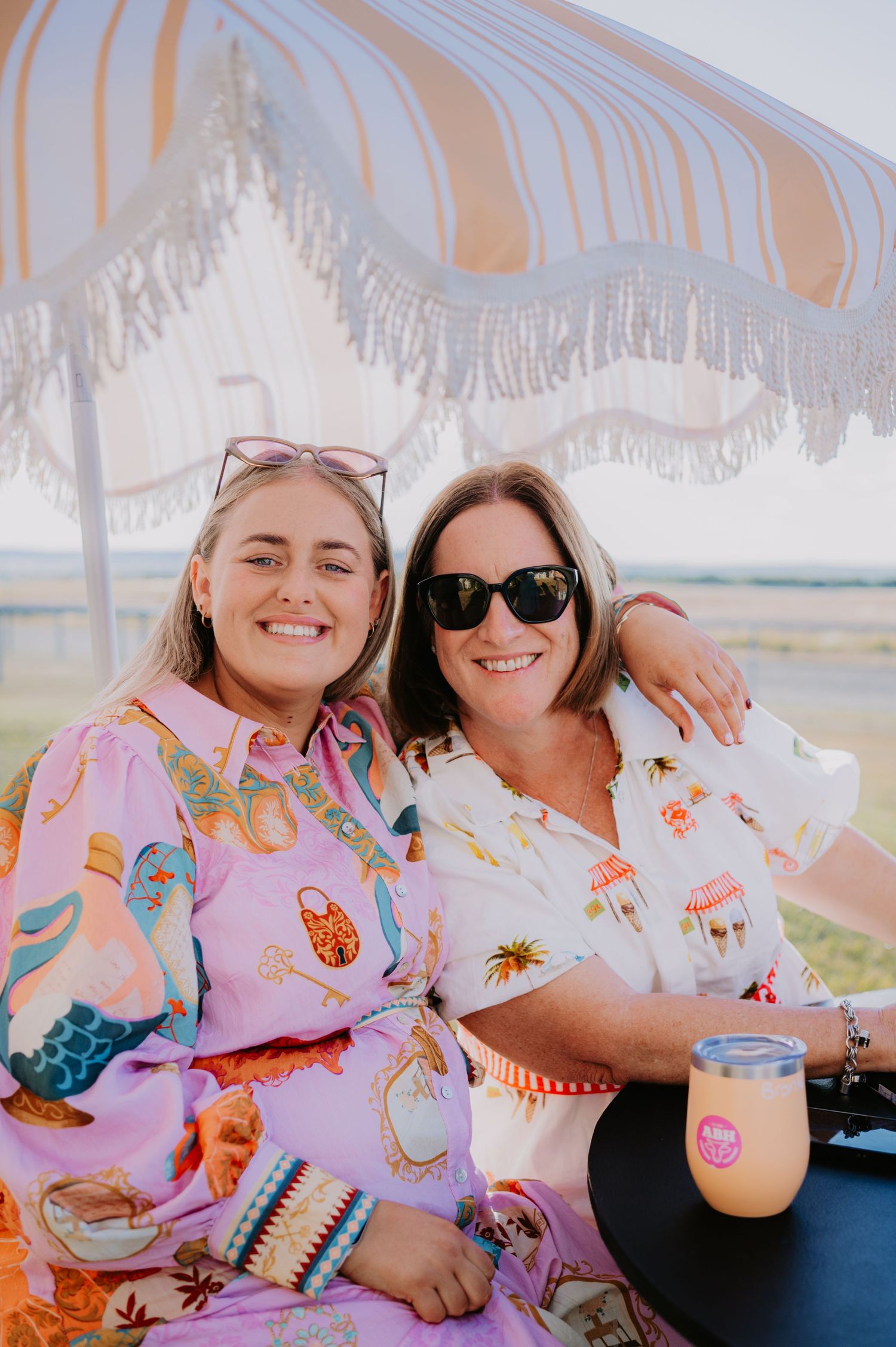 Two smiling women in colorful printed shirts pose together under a fringed beach umbrella outdoors.