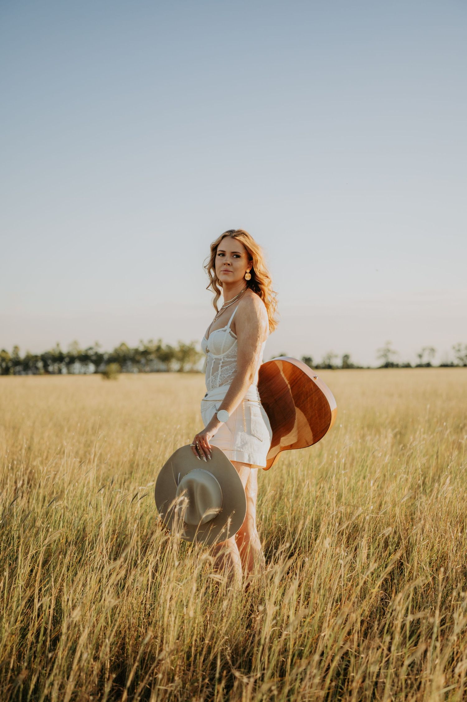 A person in a white dress twirls gracefully through a golden wheat field at sunset holding a wide-brimmed hat.