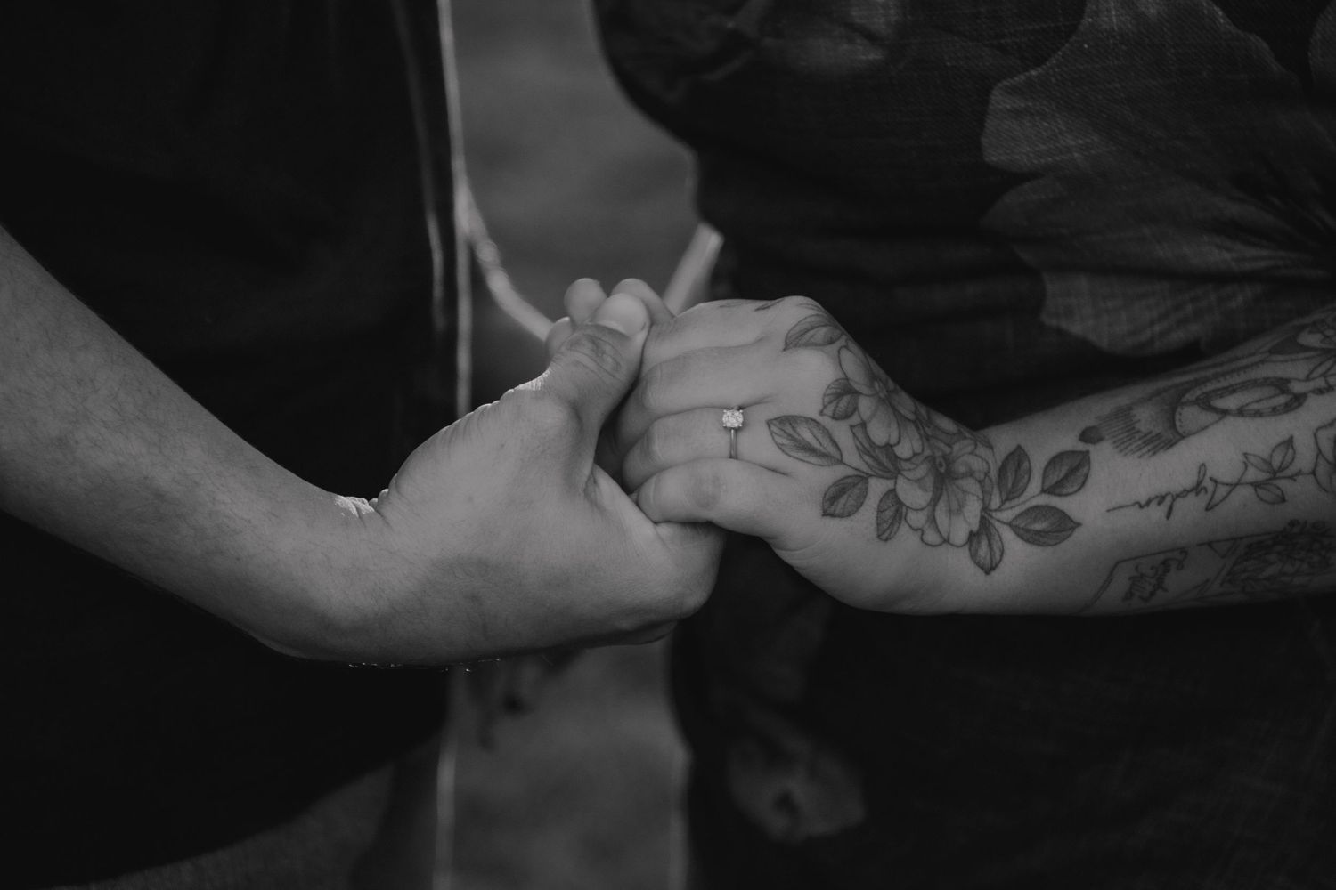 Black and white photo of two people holding hands, one with floral tattoos and a ring, symbolizing love and connection.