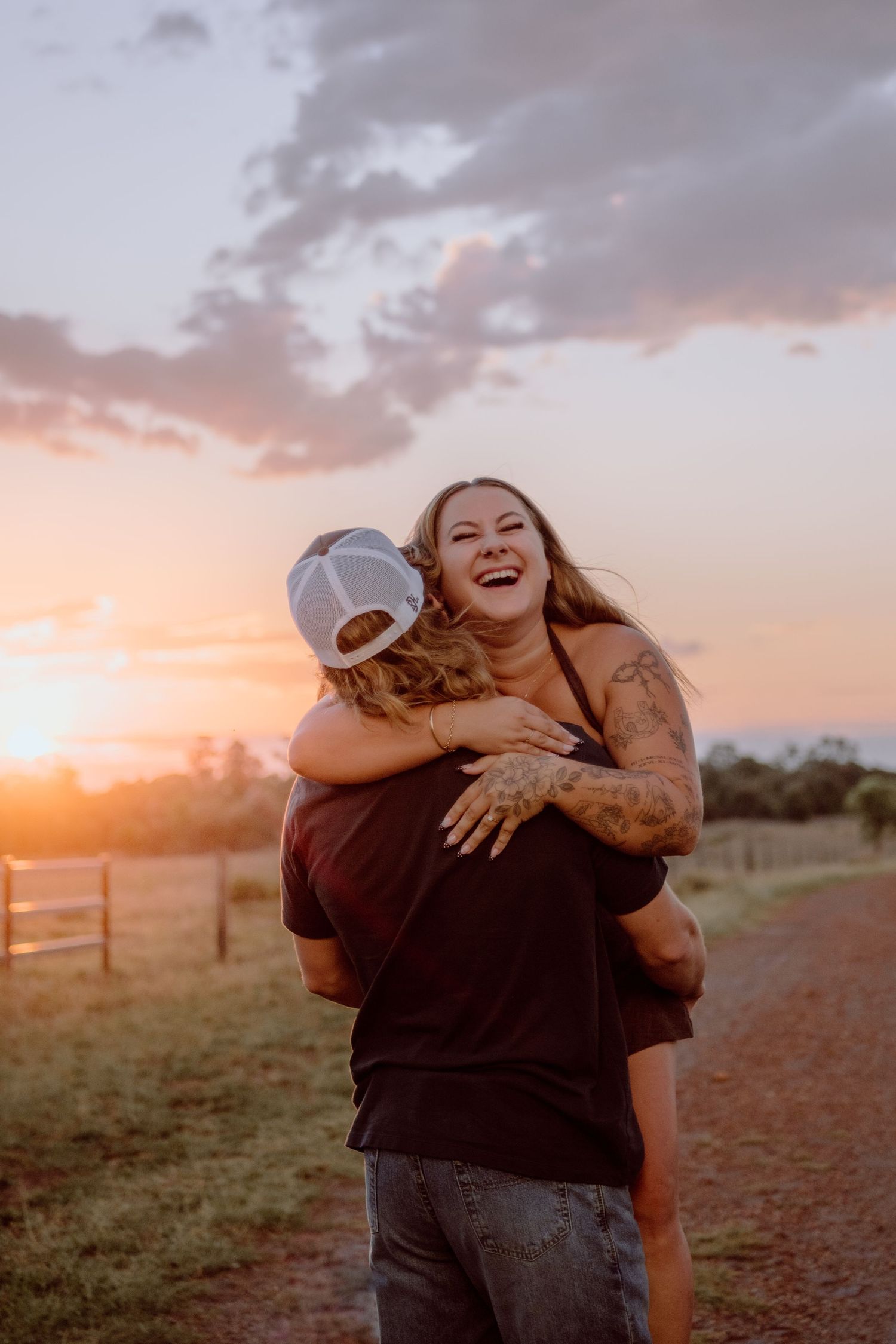 Couple embracing and laughing at sunset on a rural dirt road, woman with tattoos smiling joyfully.