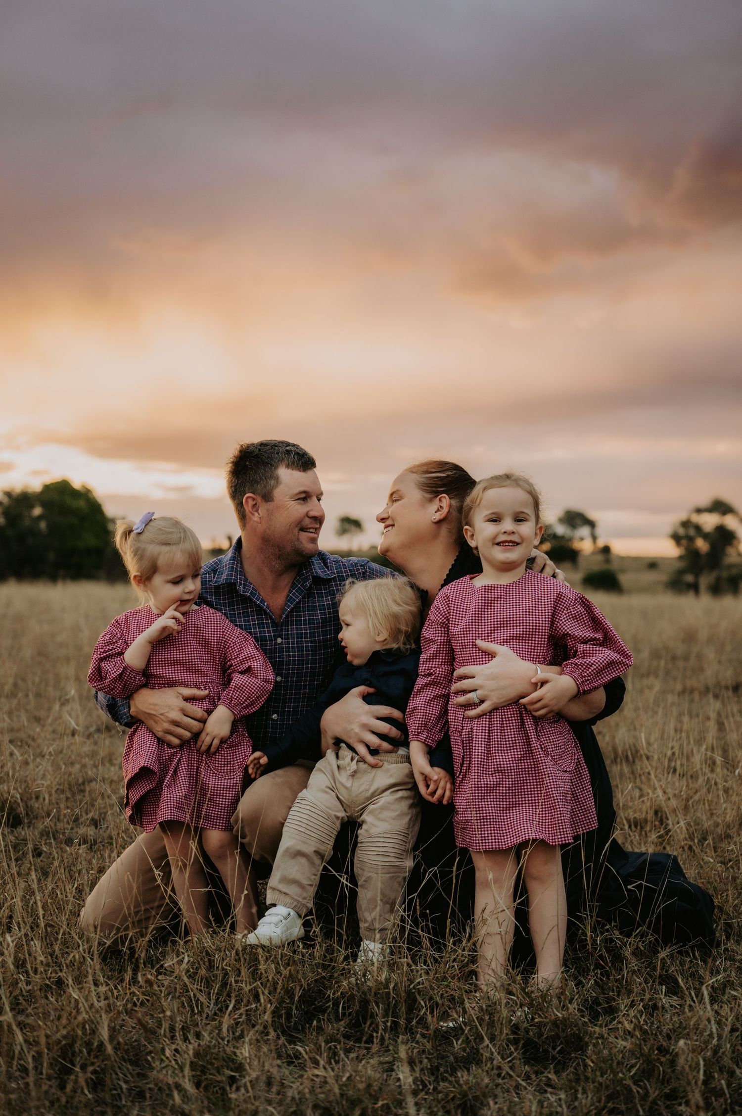 Family of five sitting in golden field at sunset, parents laughing with three young children in matching pink outfits.