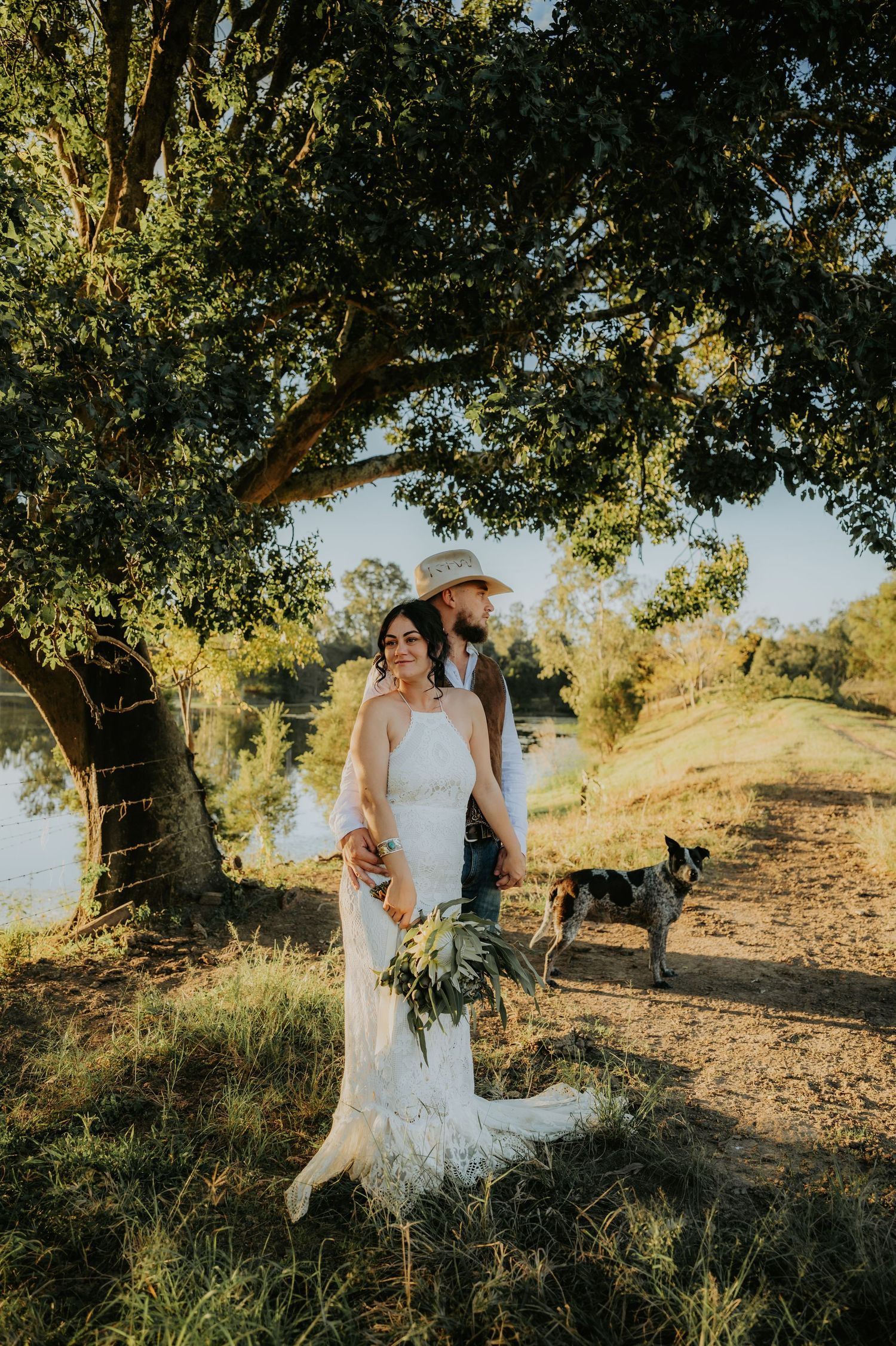 Bride in white lace gown holds bouquet with groom in cowboy hat beside a tree, dog nearby, golden hour outdoor wedding.