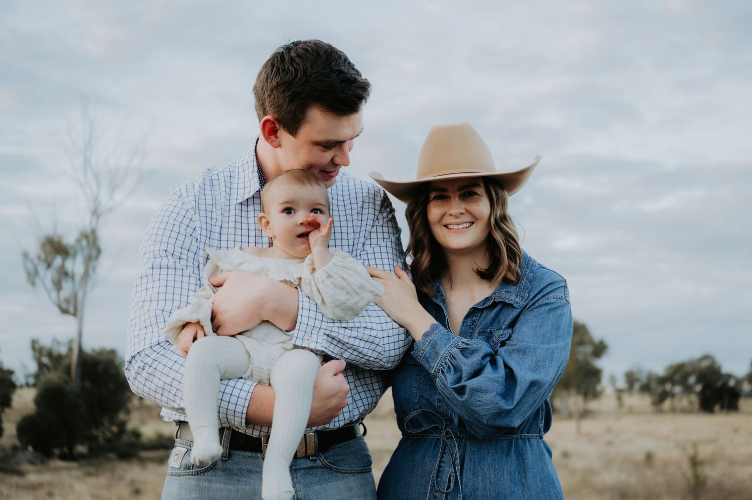 A young family with a baby poses outdoors in a rural field, mom wearing a tan hat and denim dress.