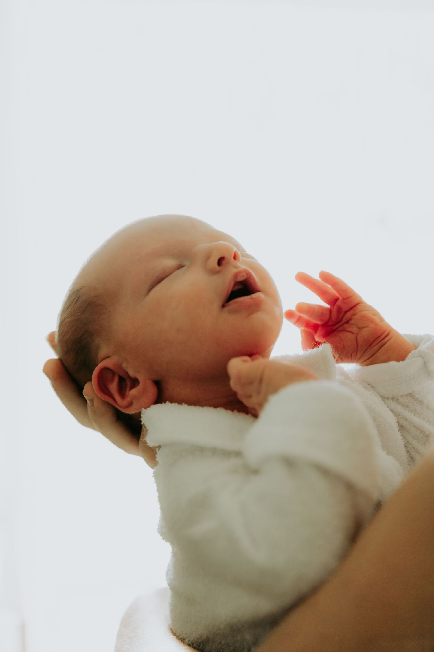 Newborn baby yawning while being held against bright background.
