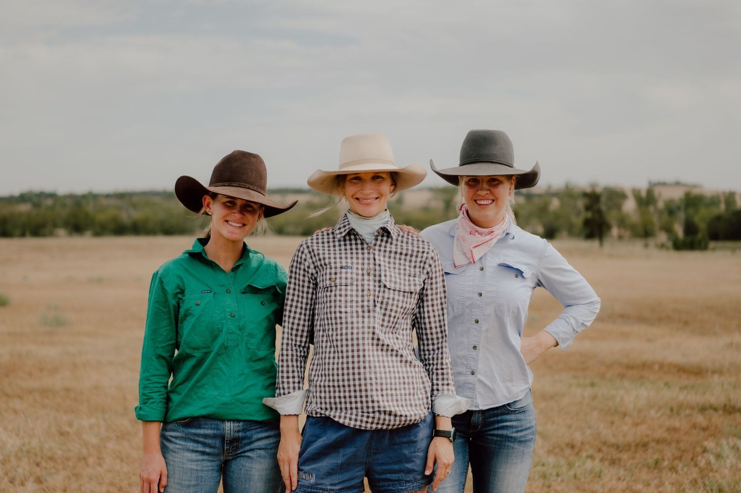 Three women in cowboy hats and western shirts smiling together in a golden field.