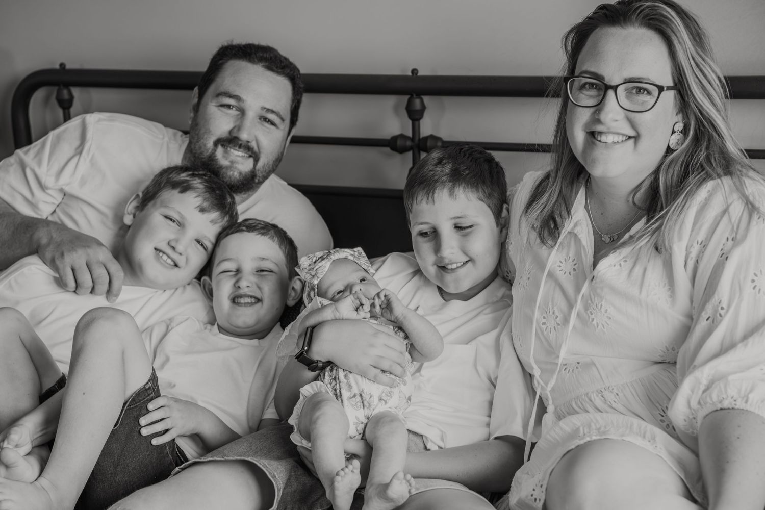 Black and white family portrait of parents with three young children and a newborn baby sitting together on a bed.
