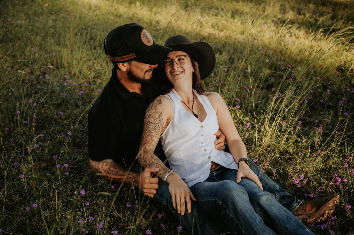 Couple sitting in a sunny meadow, woman in white tank and cowboy hat laughing, man in black shirt with tattoos.