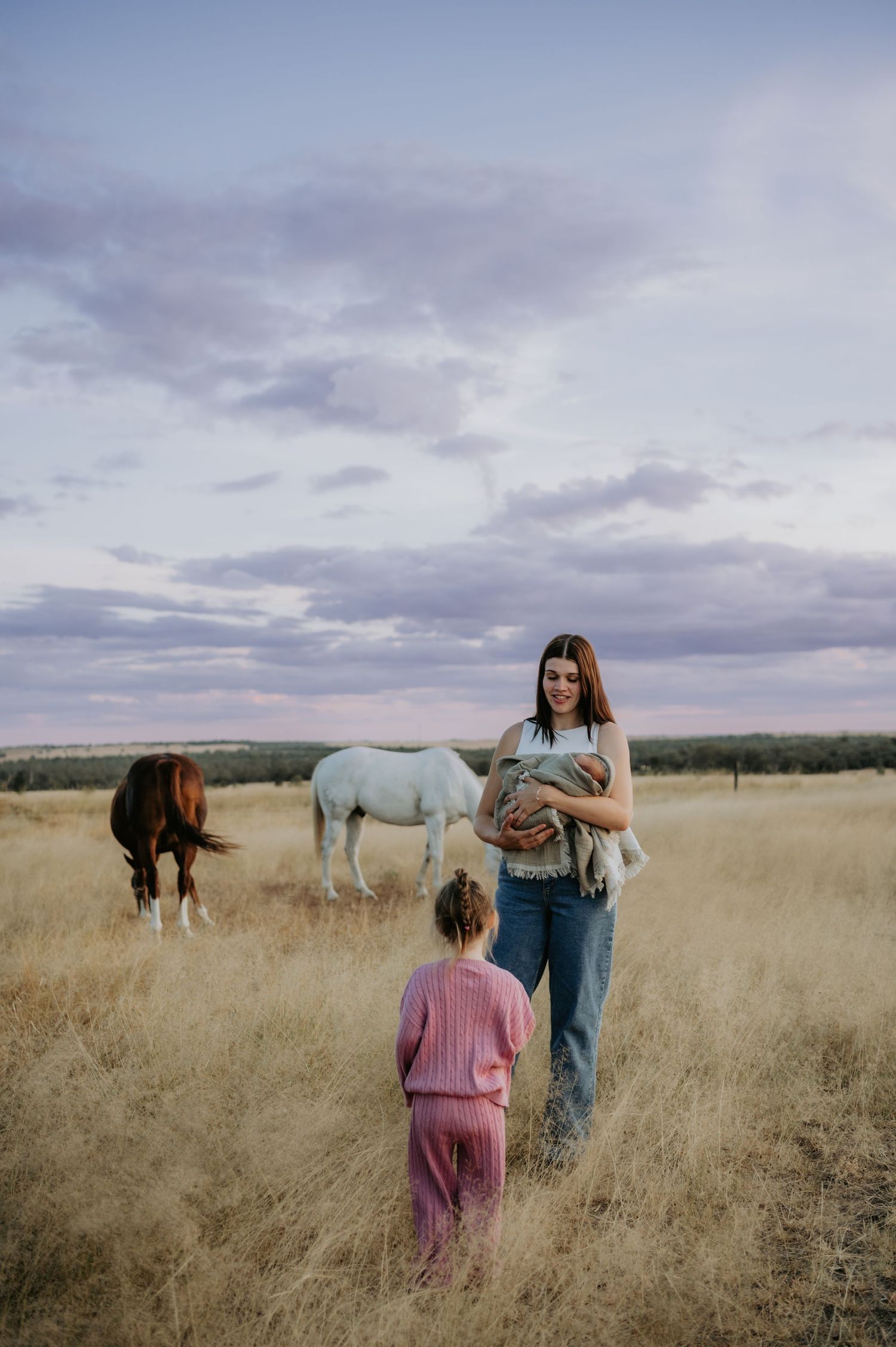 A mother holds her newborn in a golden field while her young daughter stands nearby with two horses grazing under a purple sky.