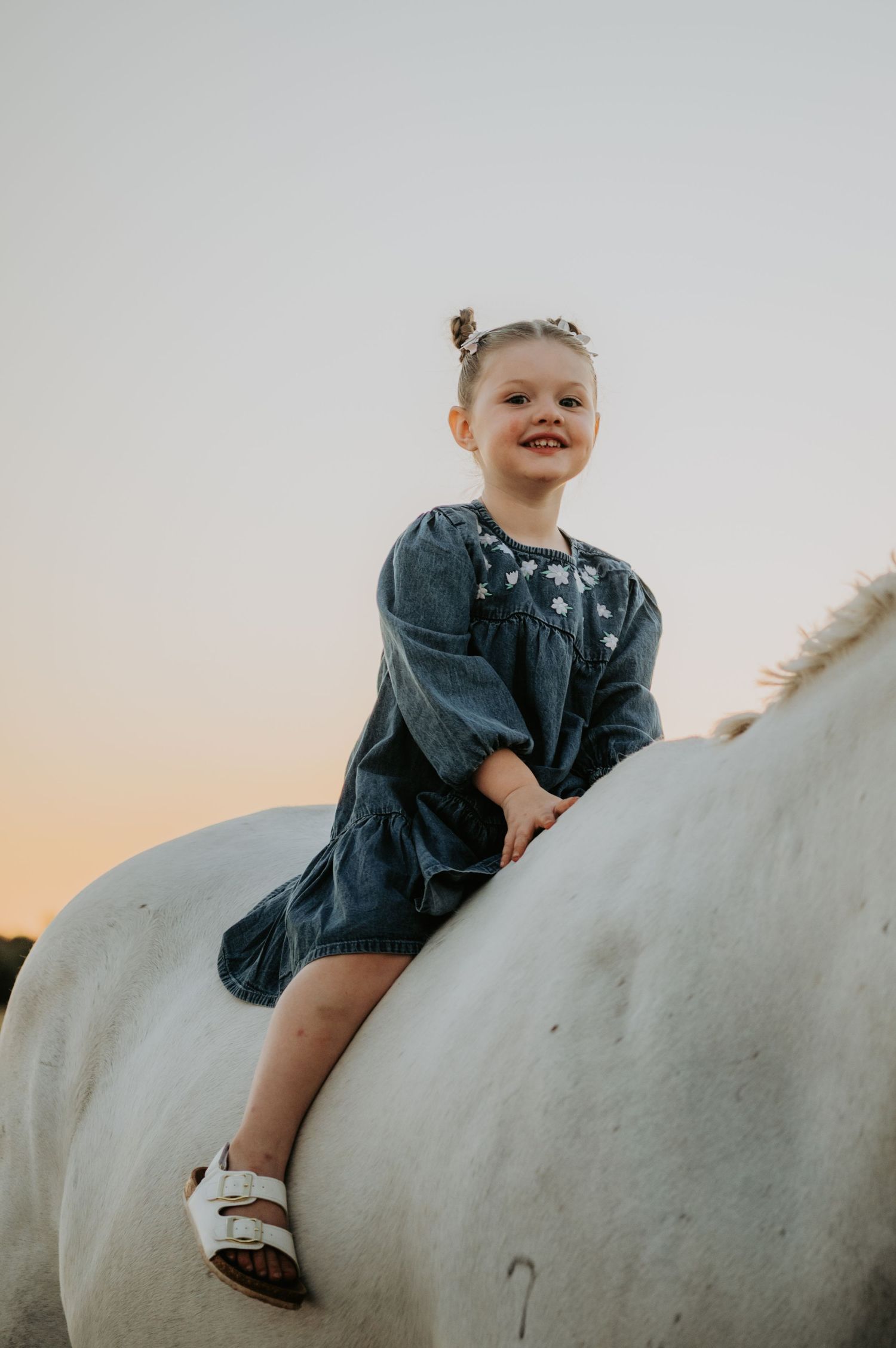 Young girl in denim dress sitting atop a white horse, smiling at sunset.