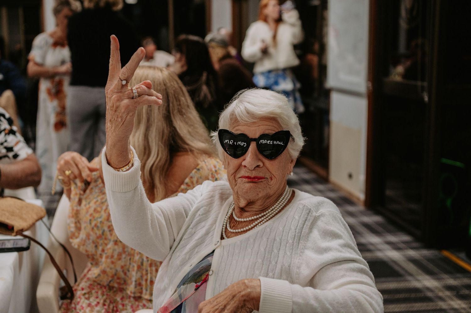 Elderly woman wearing novelty sunglasses giving rock sign at indoor event.