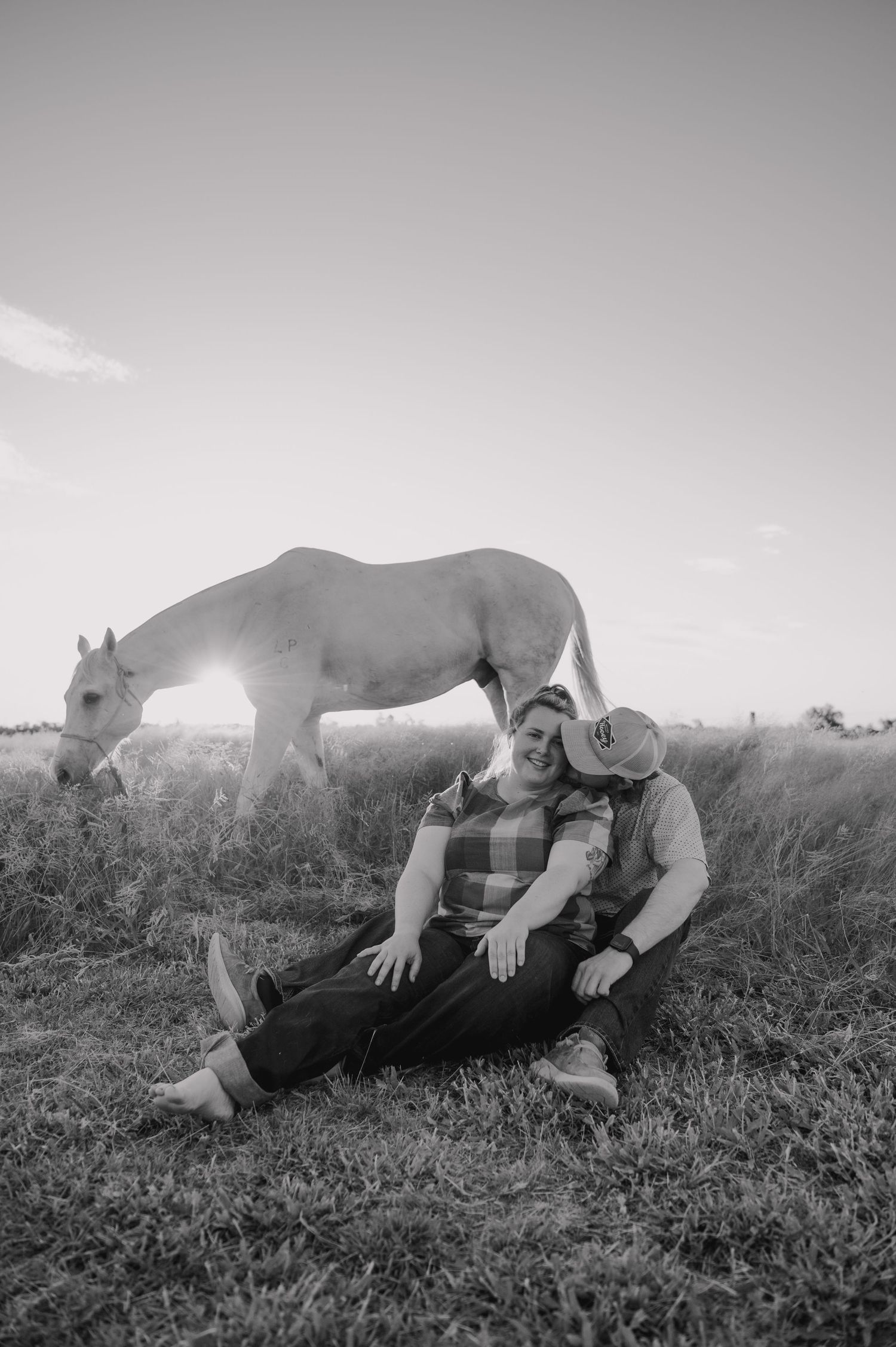 Black and white photo of couple laying in grass with horse nuzzling them.