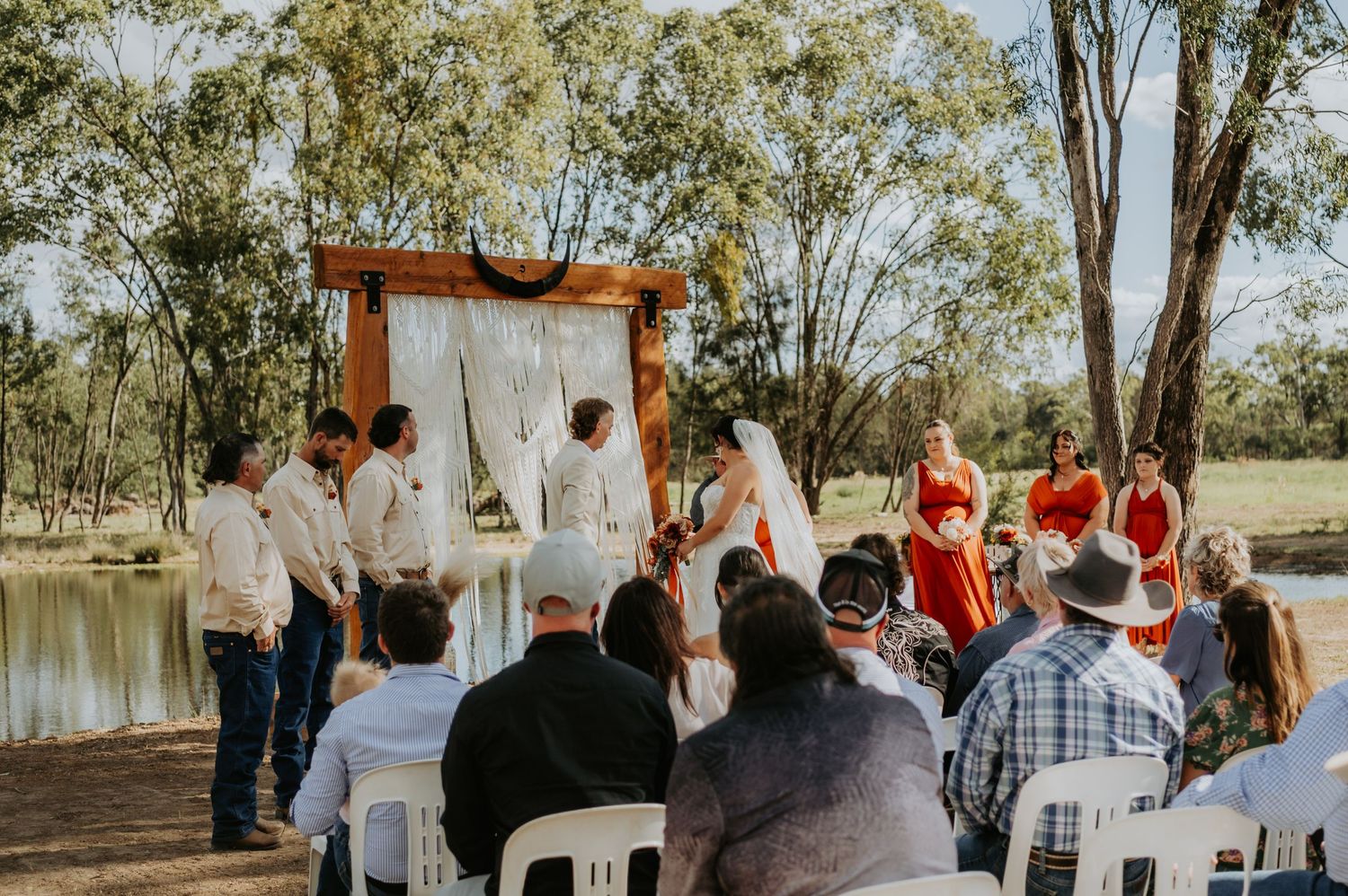Outdoor wedding ceremony by a pond with guests seated in white chairs beneath a rustic wooden arch.