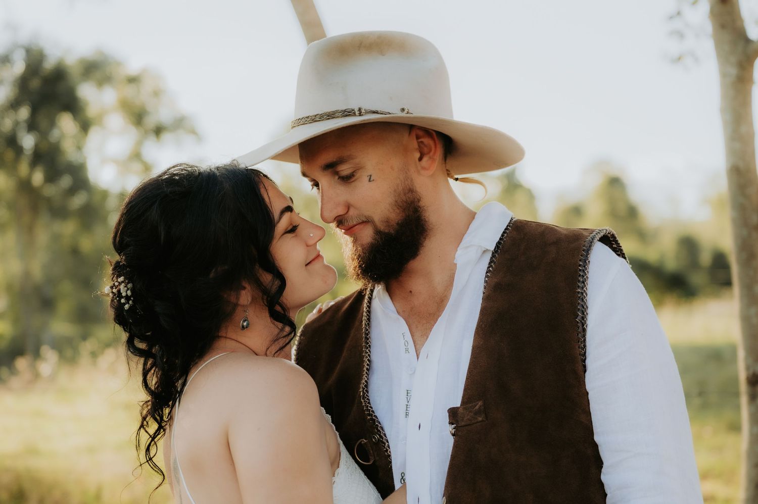 Romantic couple in boho wedding attire sharing tender moment outdoors, groom in wide-brim hat and vest, bride with dark updo.