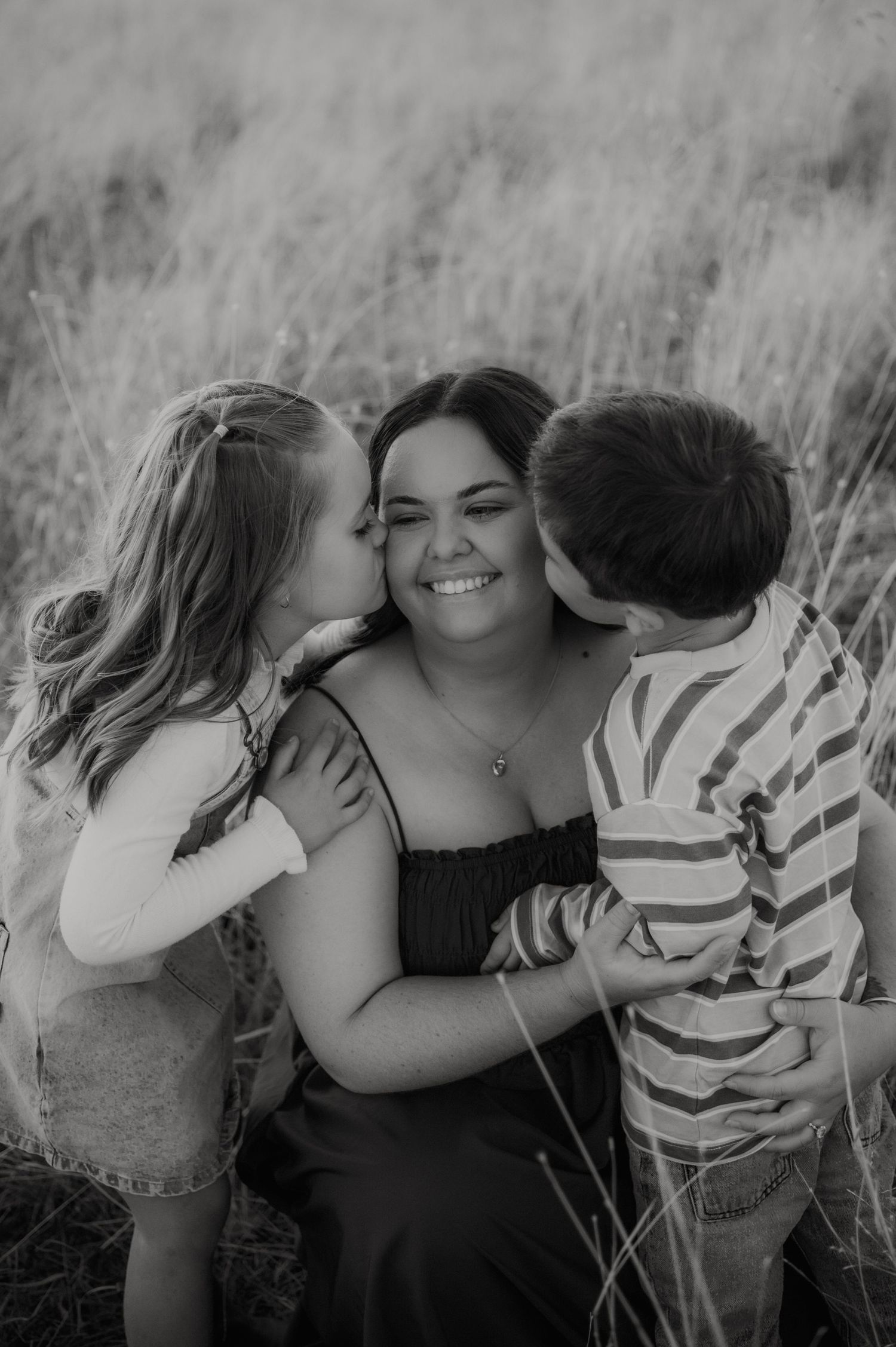 Black and white photo of a smiling woman being kissed on both cheeks by two young children in a grassy field.
