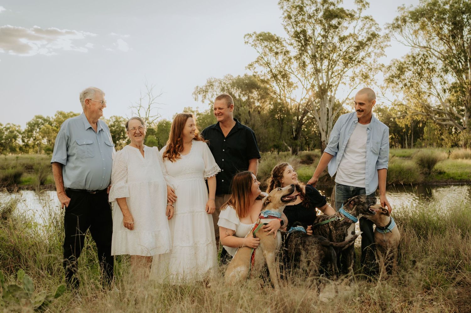 Multi-generational family gathering outdoors near a pond with dogs, surrounded by trees at golden hour.