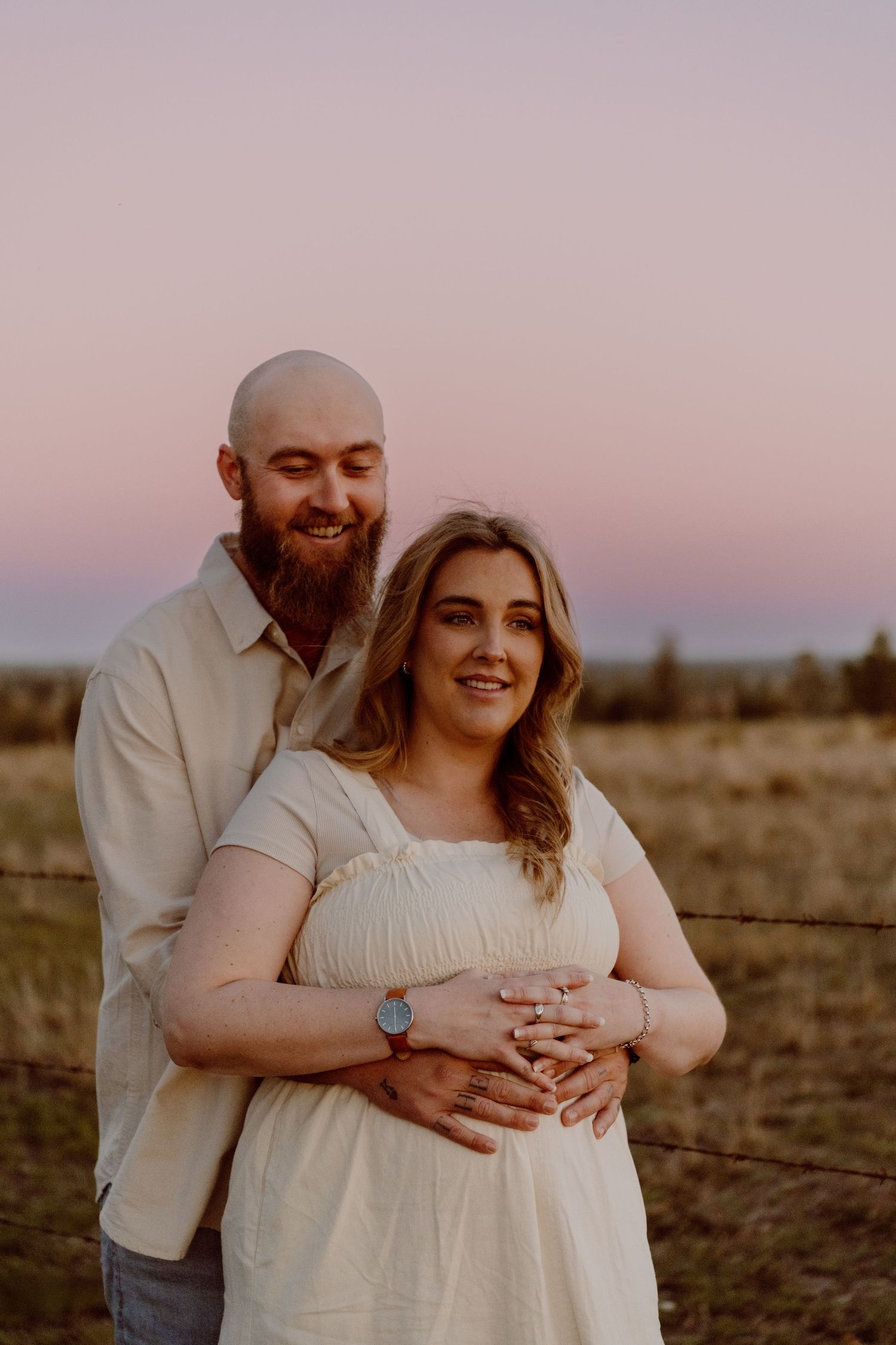 A couple poses at sunset in a field, man embracing pregnant woman in white dress, both smiling warmly.
