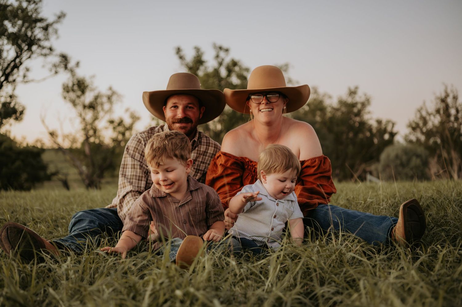 Family of four in cowboy hats sitting in green grass at golden hour, smiling outdoors.