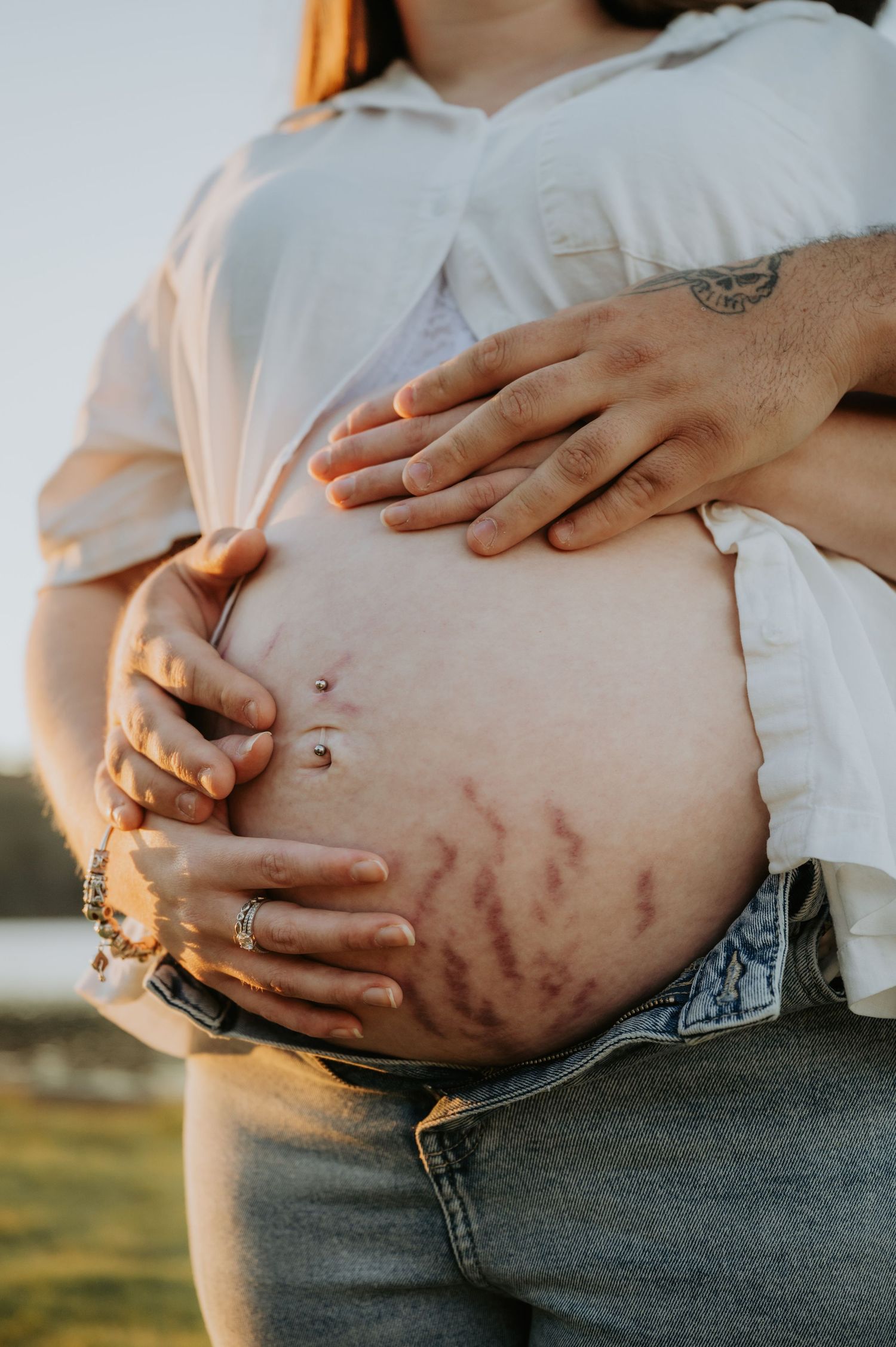 Pregnant woman's bare belly with stretch marks and belly piercing, cradled by two pairs of hands outdoors.