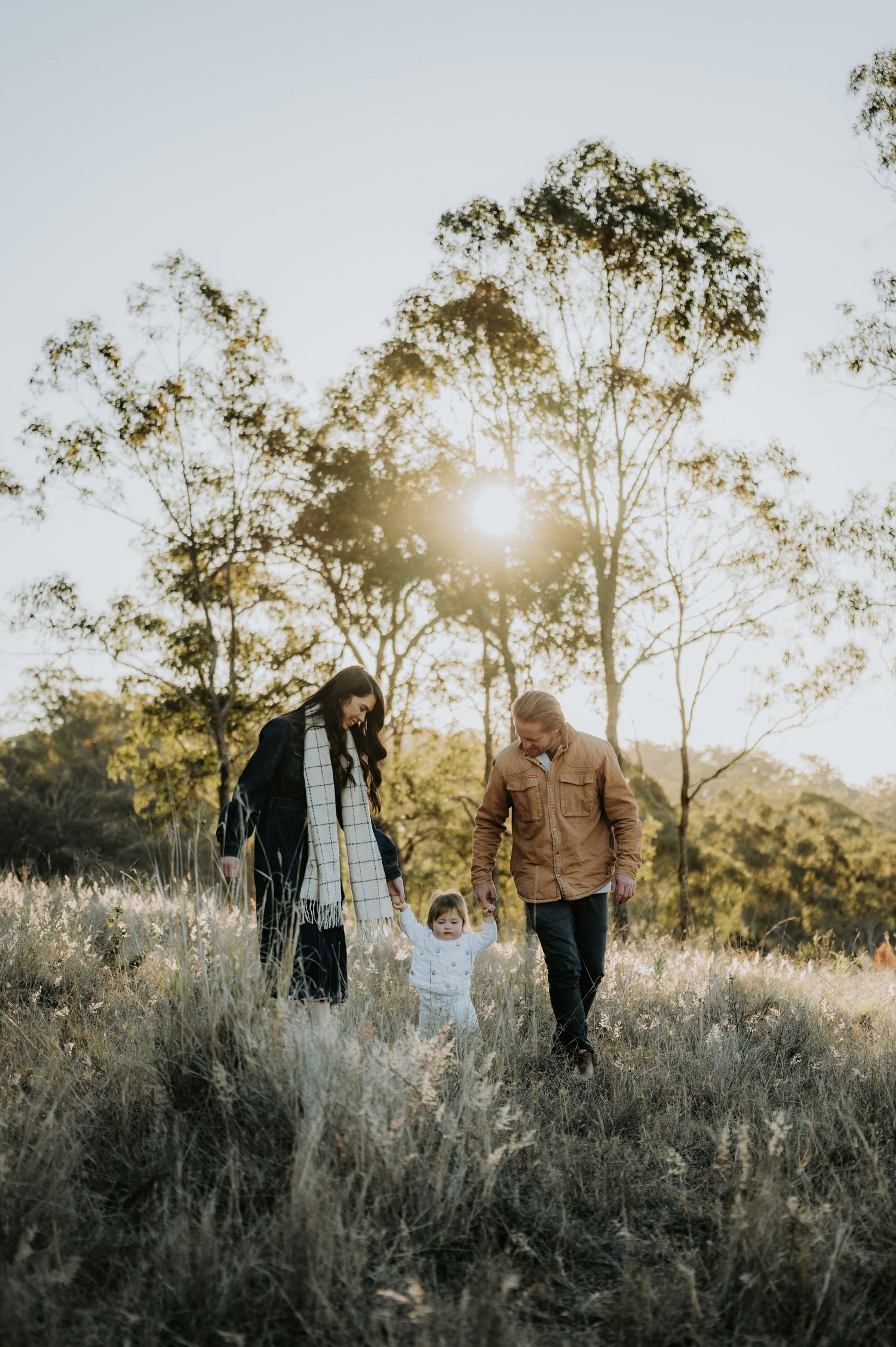 A family of three walks through a sunlit field, parents holding toddler's hands, surrounded by tall grass and eucalyptus trees.