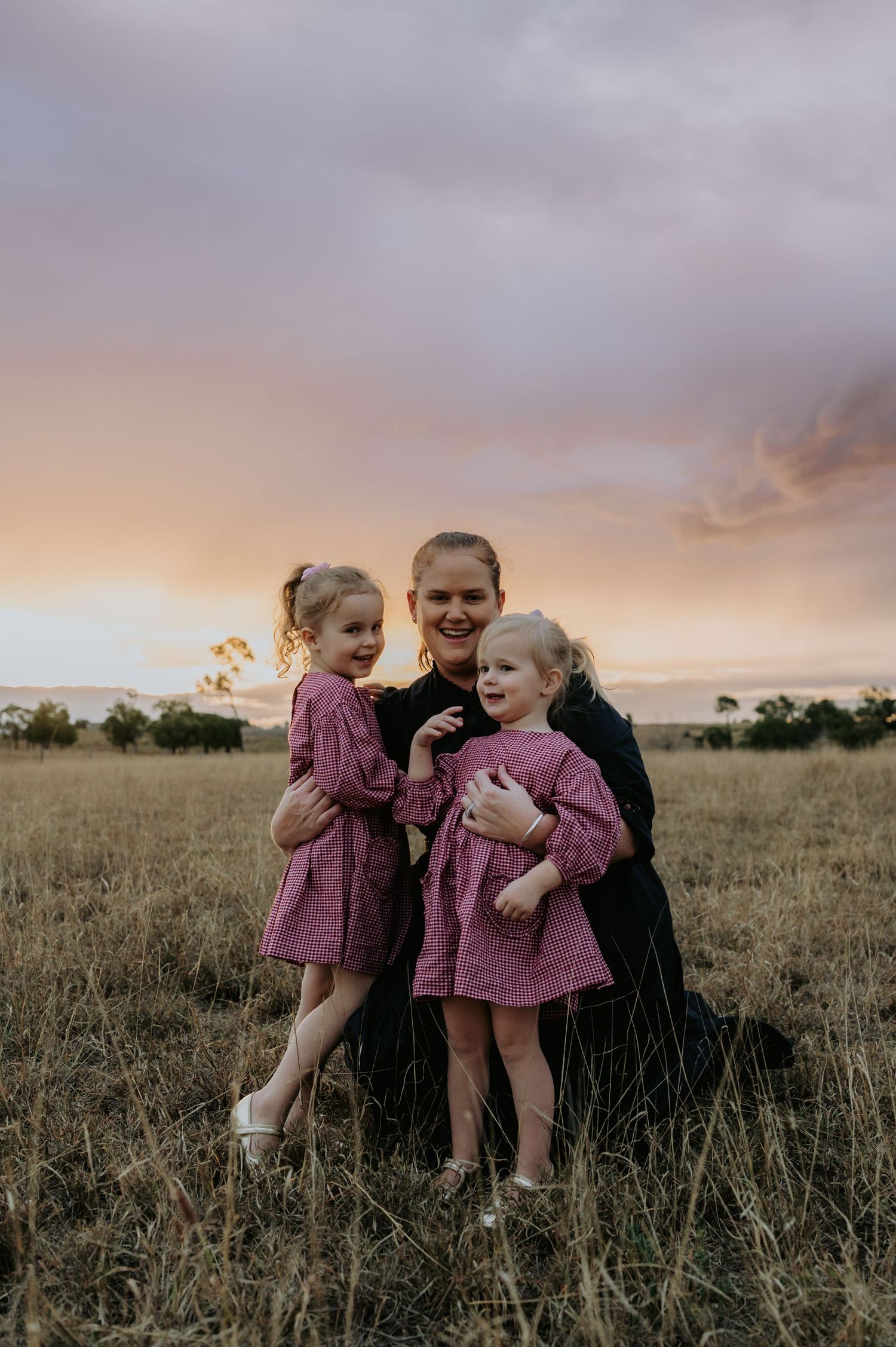 A mother hugs her two young daughters in pink dresses in a grassy field at sunset.