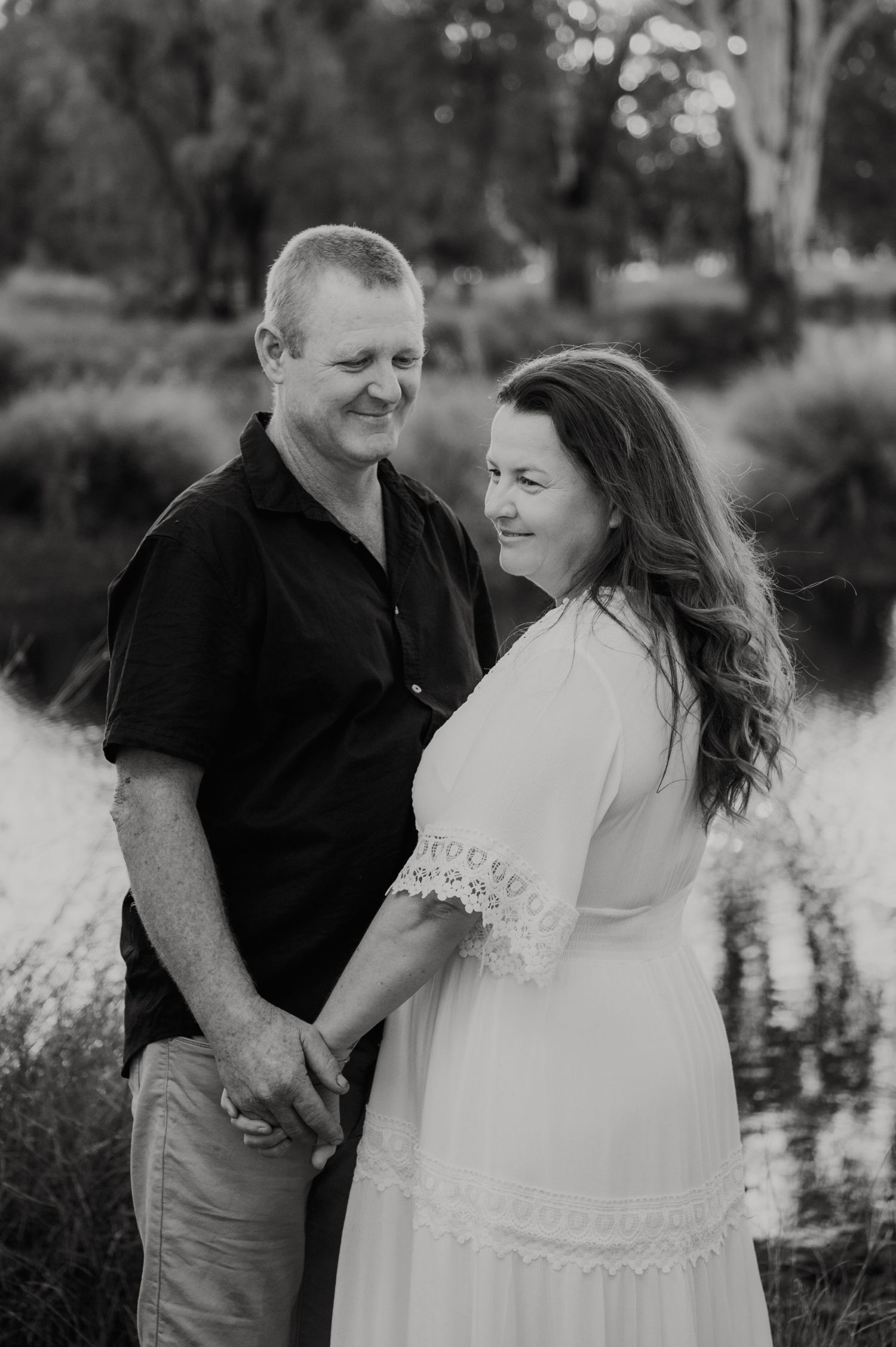 Black and white photo of a couple holding hands and smiling at each other outdoors near a pond.